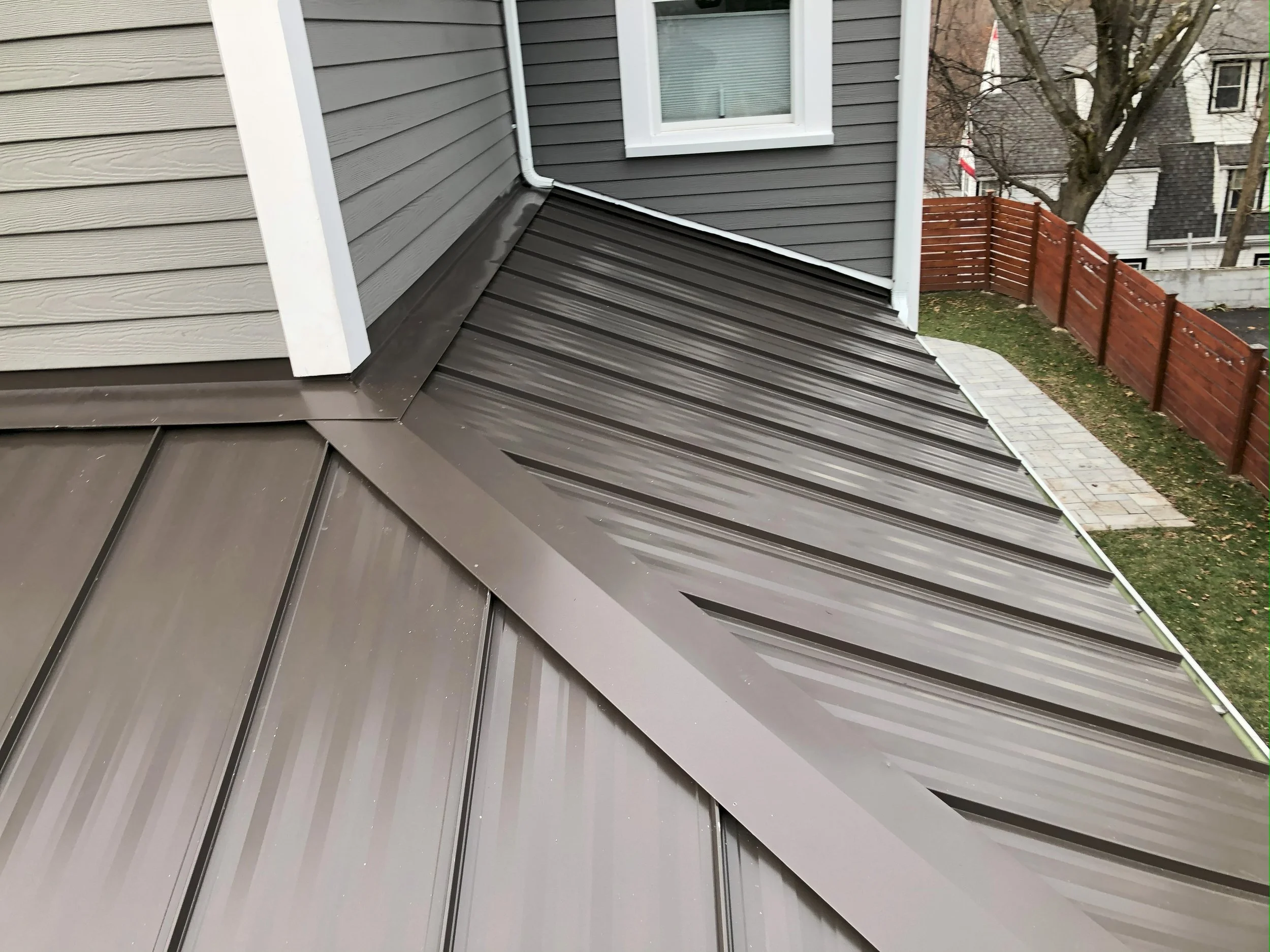 View of a metal roof with brown panels, corner intersection with a house's gray siding, a window, and a wooden fence in the background.