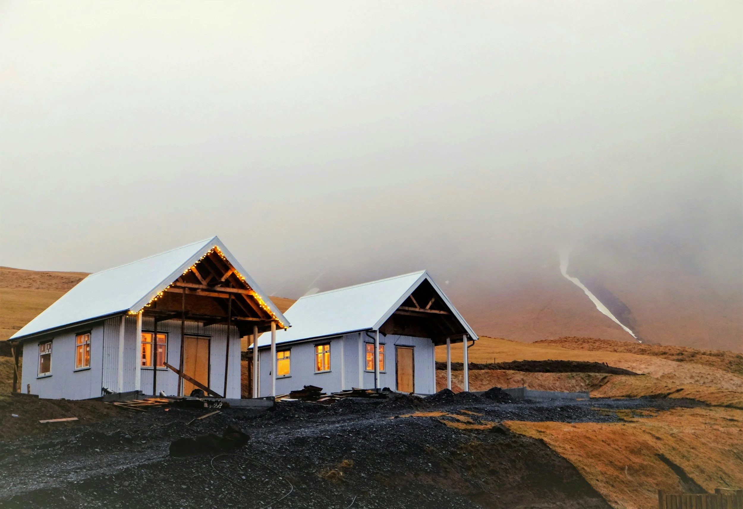 Two small houses under construction on a hillside at sunset, with mountains and waterfalls in the background.