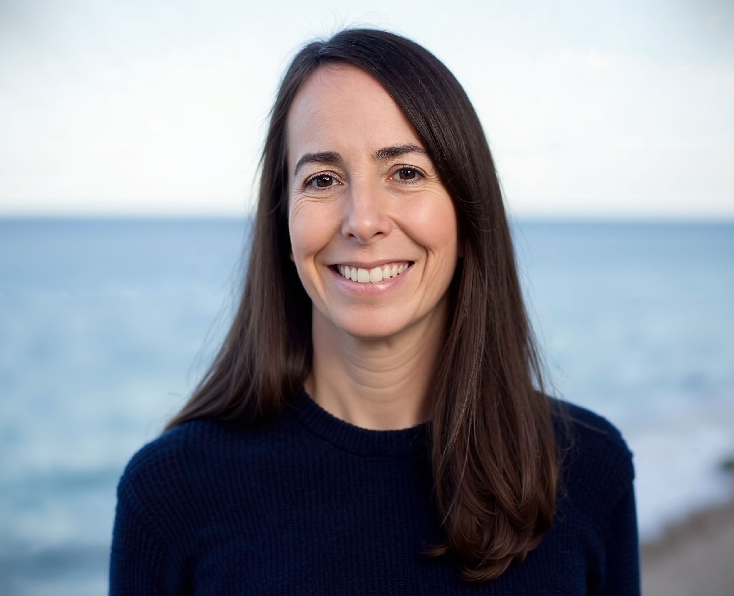A woman with long brown hair smiling outdoors near a body of water.