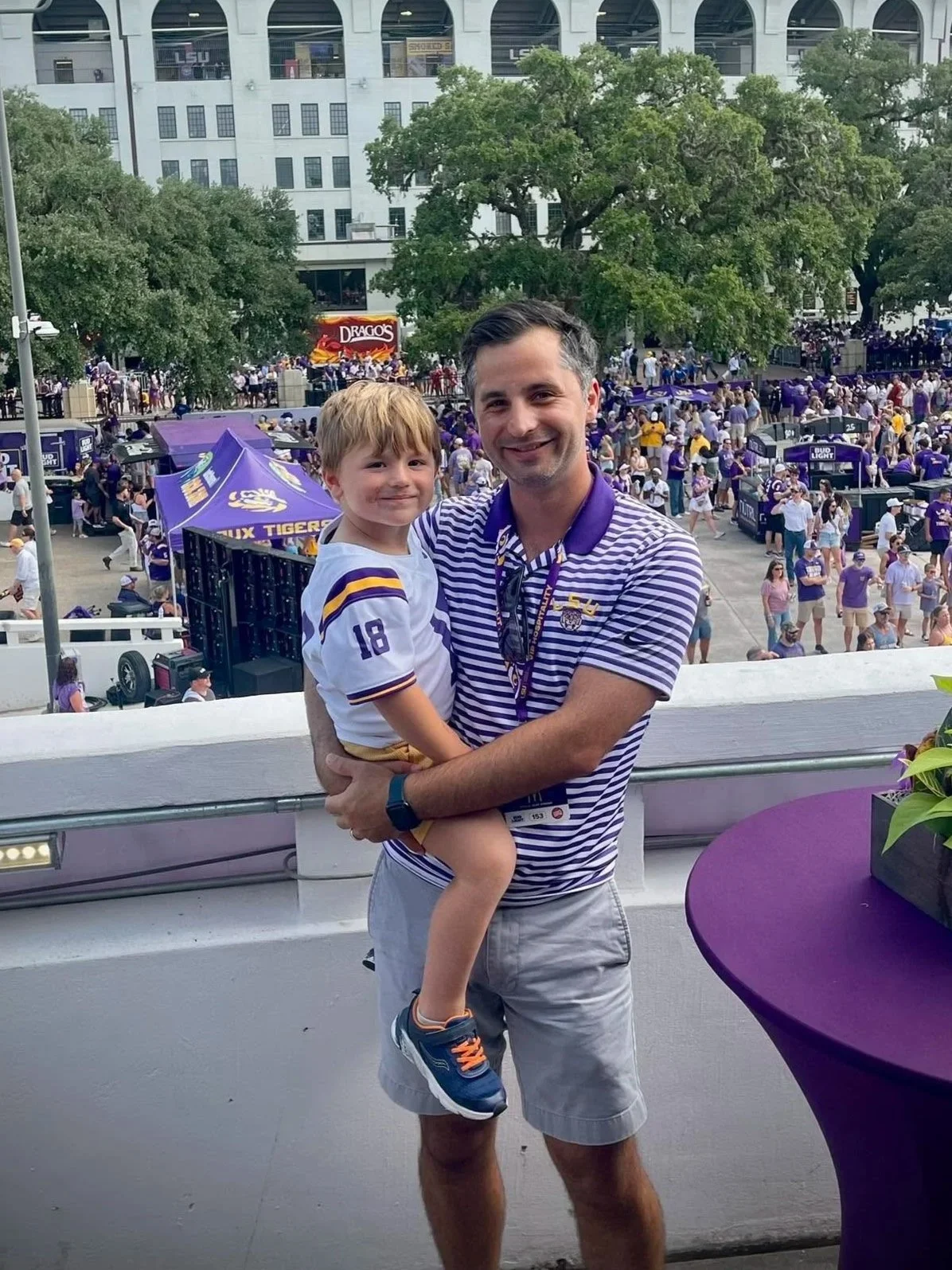 A smiling man holding a young boy in his arms on a balcony, with a crowd gathered in the background at a sports stadium or arena, showing purple decorations and tents.