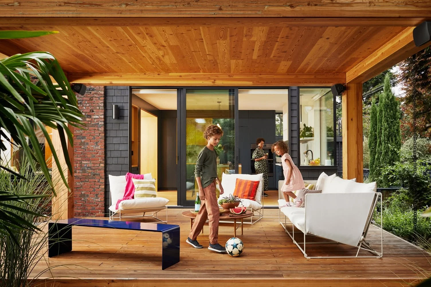 Children playing with a soccer ball on an outdoor wooden patio furnished with white and colored cushions, with a view into a modern kitchen and garden.