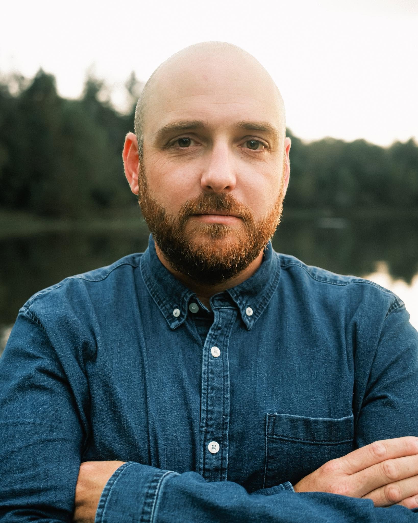 Close-up of a man with a shaved head and reddish beard wearing a denim shirt, standing outdoors with a blurred background of trees.