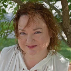 A woman with curly, reddish hair smiling outdoors with trees in the background.