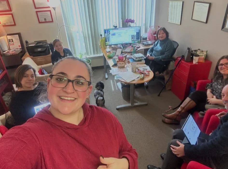 Group of six women and a small dog in an office, with one woman taking a selfie. The room has a desk with a computer, papers, and snacks, and framed certificates on the wall.