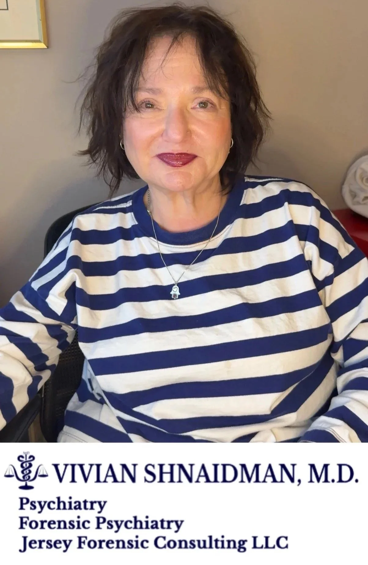 A woman with short, wavy dark hair wearing a blue and white striped shirt, sitting indoors, with a business card displaying her name Vivian Shaidman, M.D., and her specialties in psychiatry, forensic psychiatry, and consulting.