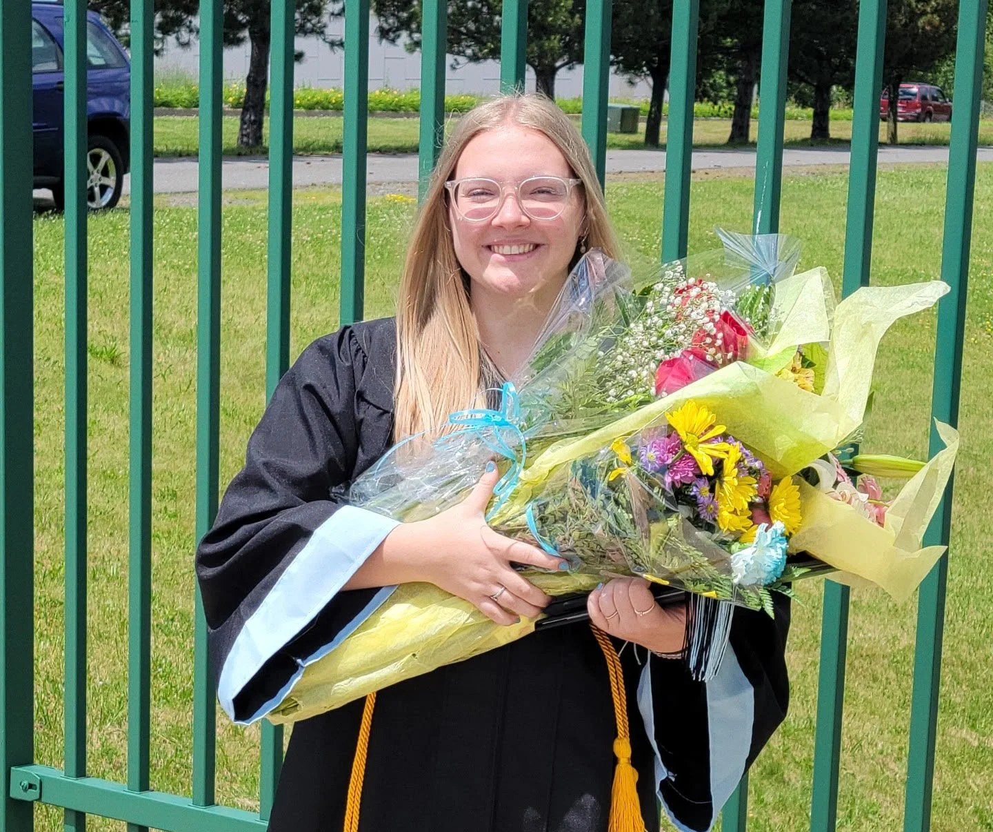 A young woman in a burgundy graduation gown holding a bouquet of flowers, smiling at night in front of lit palm trees and water fountains at an outdoor shopping center or plaza.