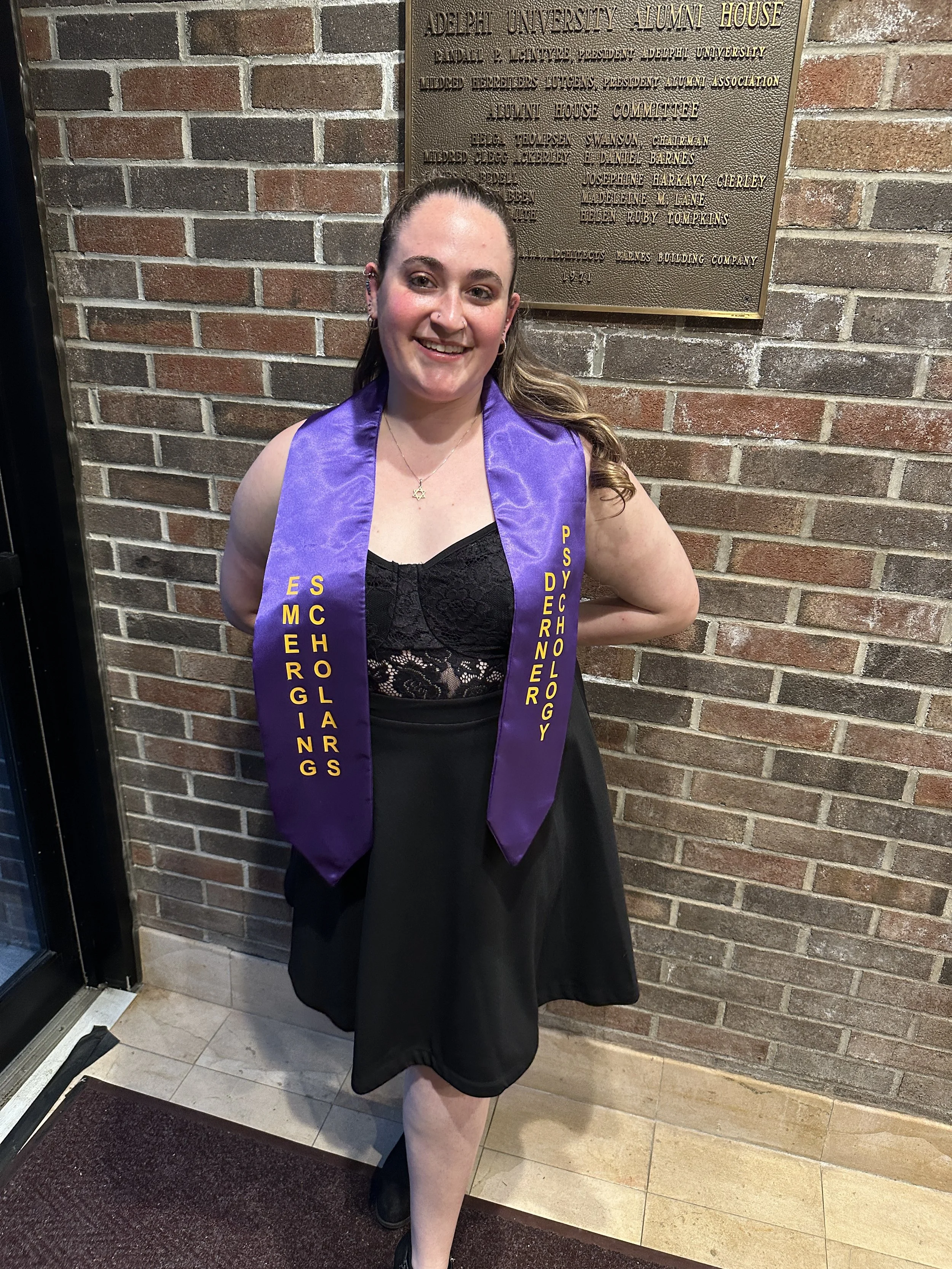 A smiling woman in a black dress standing in front of a brick wall with a bronze plaque, wearing a purple graduation stole that says 'Emerging Scholargs Psychology'.