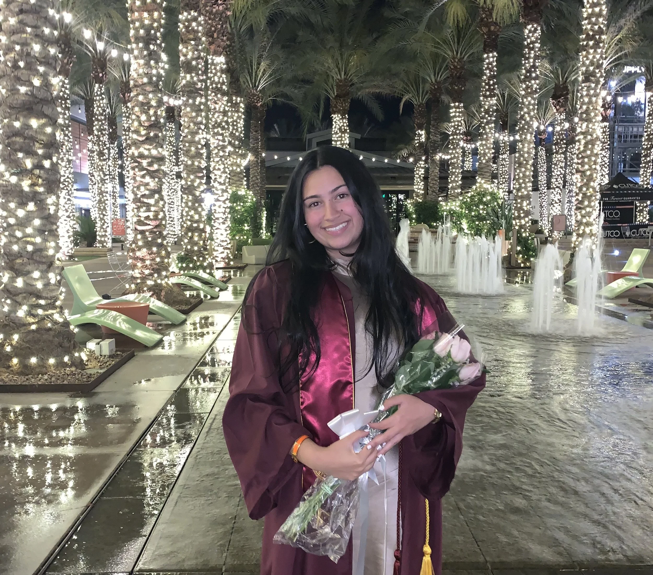 A young woman in a maroon graduation gown holding a bouquet of pink roses stands outdoors at night, in front of a backdrop of palm trees decorated with string lights and a fountain.