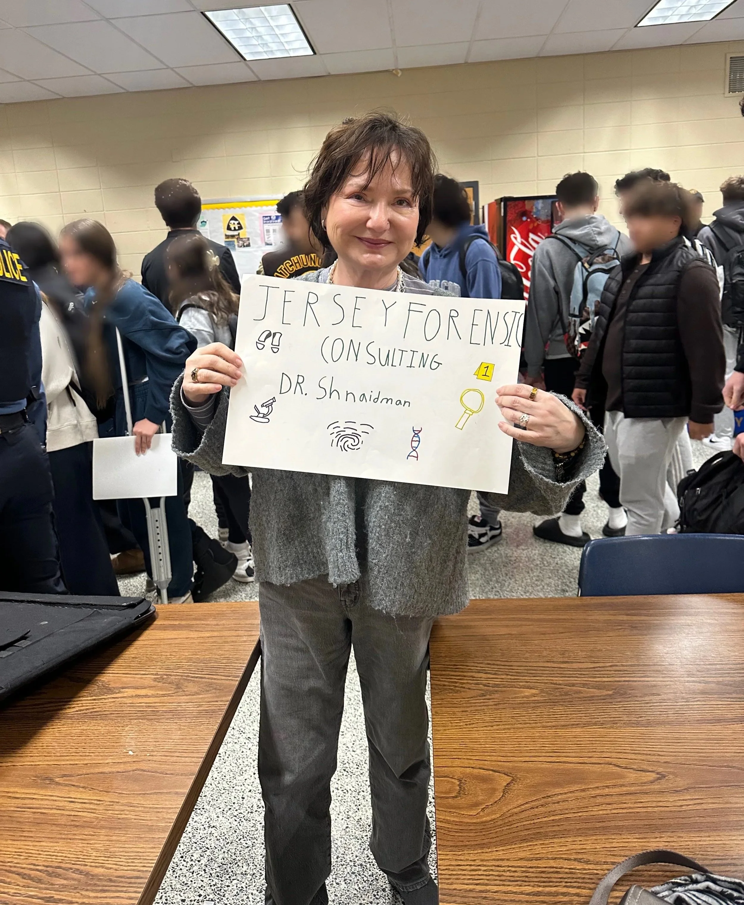 A woman holding a sign that reads 'Jersey Forensic Consulting Dr. Shnaidman' with symbols for science, fingerprint, DNA, magnifying glass, and microscope, in a school hallway with students and police in the background.