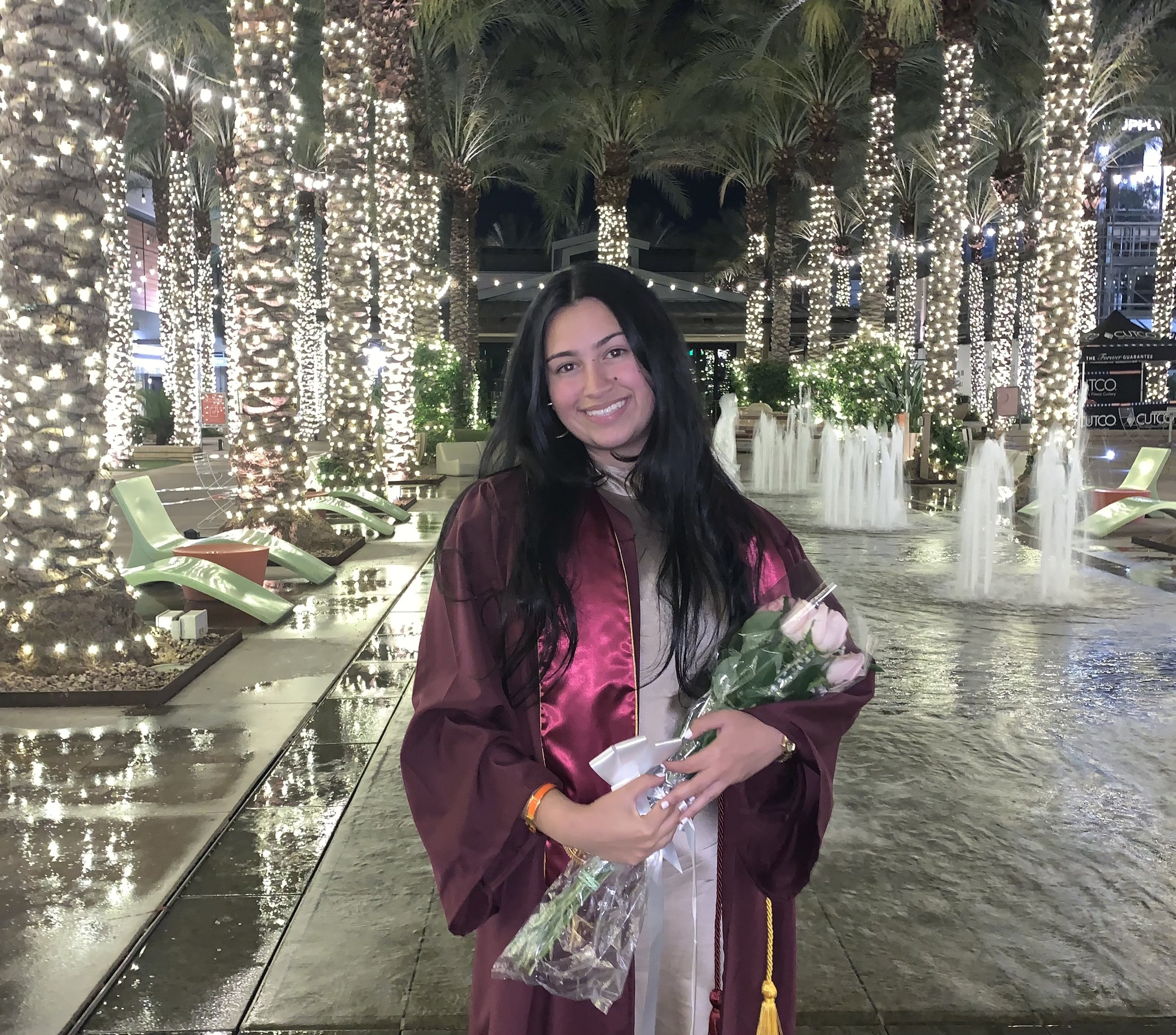 A young woman in a burgundy graduation gown holding a bouquet of flowers, smiling at night in front of lit palm trees and water fountains at an outdoor shopping center or plaza.