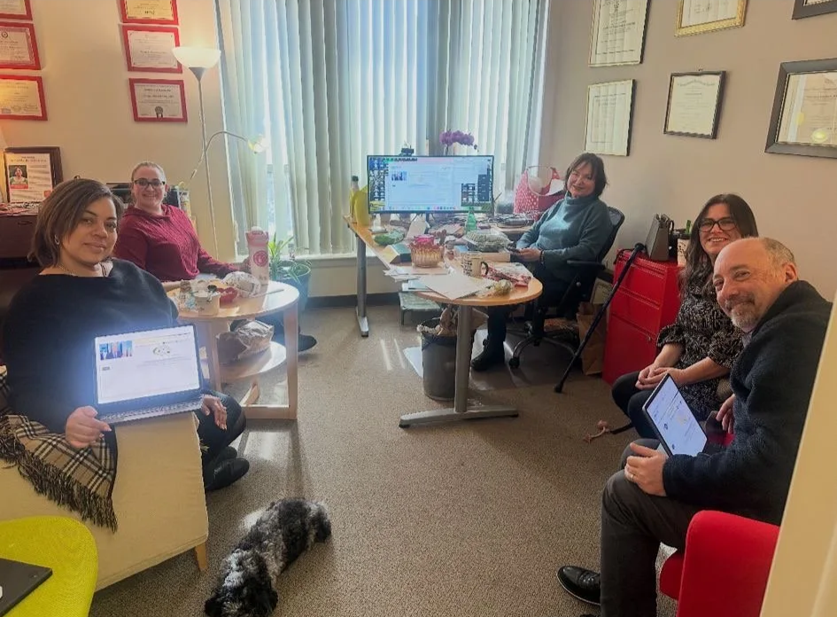 Five people sitting in an office with work desks and computers, four women and one man, smiling at the camera, with certificates or awards on the wall, and a dog lying on the carpet in the foreground.