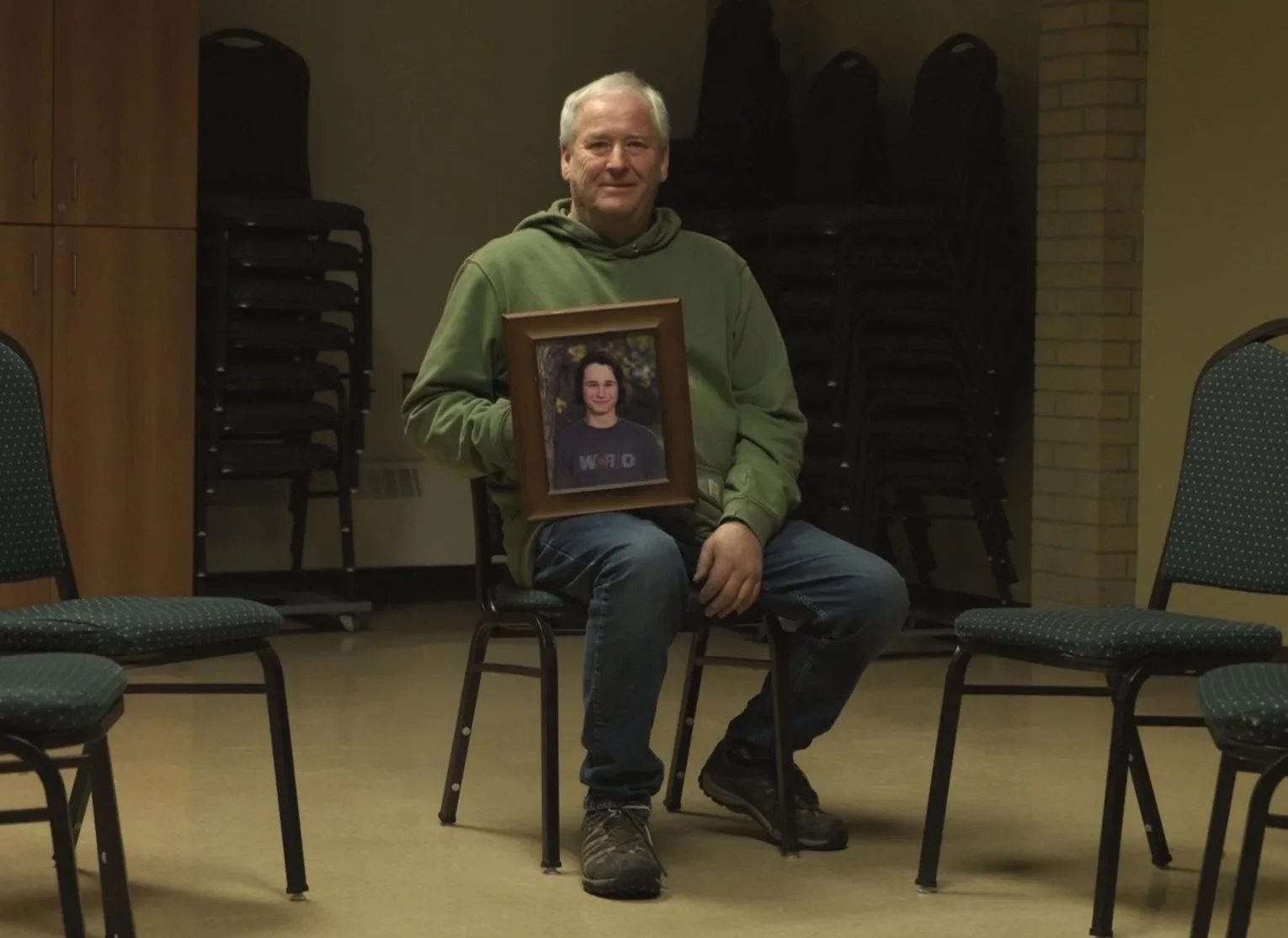 A man sitting on a chair in a room holding a framed photograph of a young man smiling outdoors. The room has stacked chairs, a wooden cabinet, and a brick wall.