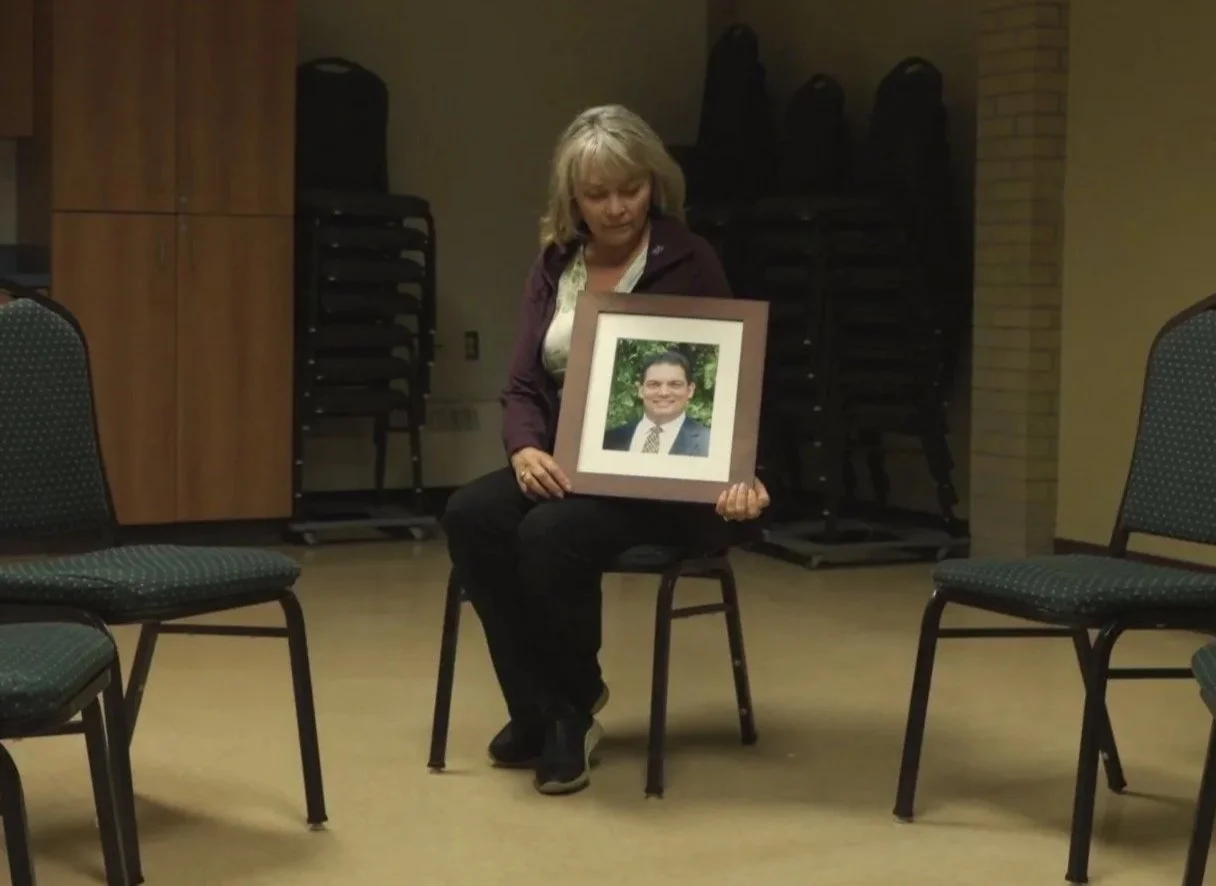 Woman sitting on a chair in a room with empty chairs, holding a framed photograph of a smiling man in a suit and tie.