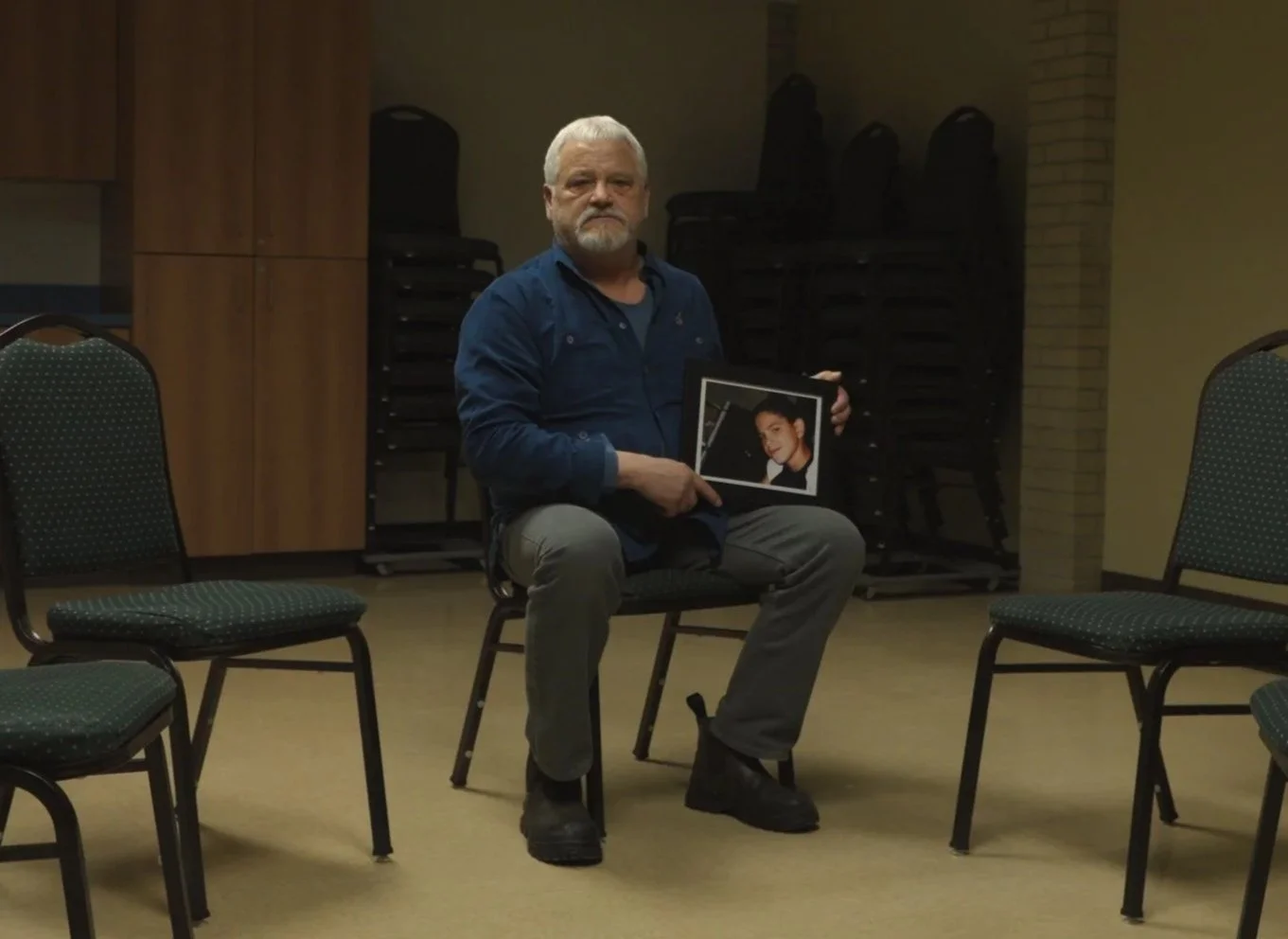 A man with white hair and a beard sits on a chair in an empty room, holding a framed photograph of a young man.