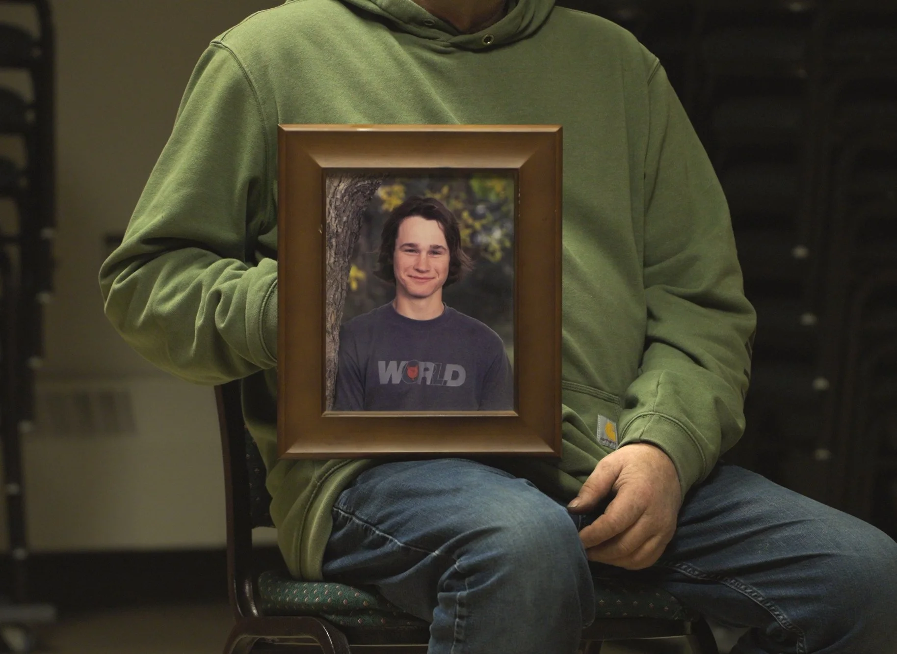 Person in green hoodie holding a framed photograph of a smiling young man with shoulder-length dark hair standing outdoors near a tree.