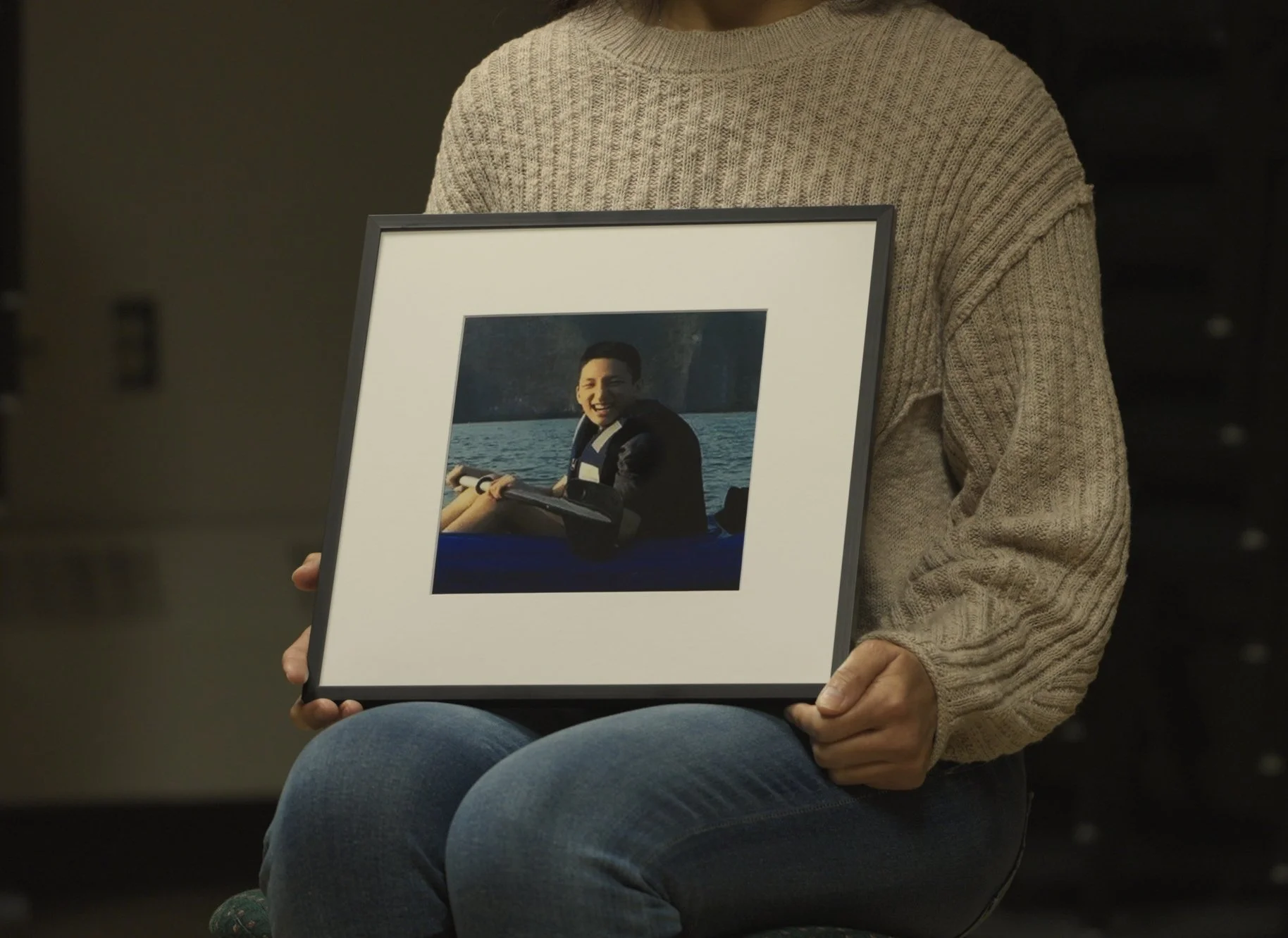 Person in a beige sweater holding a framed photograph of a smiling person in a life jacket on a boat, with water and mountains in the background.