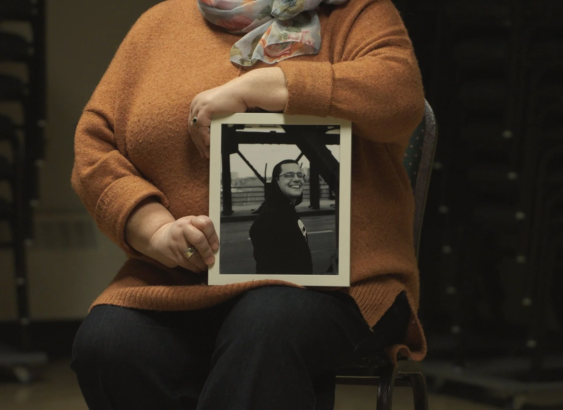 A person wearing an orange sweater and a patterned scarf holding a framed photograph of a smiling woman with glasses outdoors, with a bridge structure in the background.