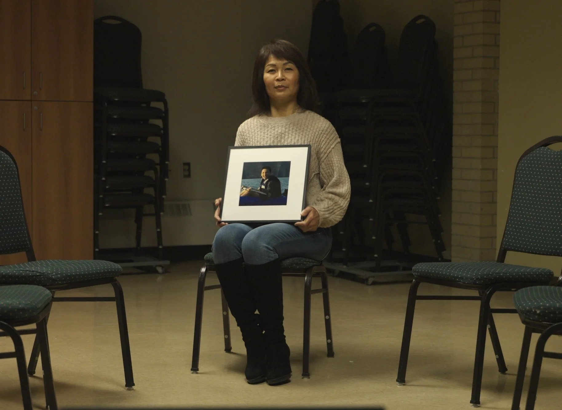Woman sitting on a chair in a room, holding a framed photograph of a young man.