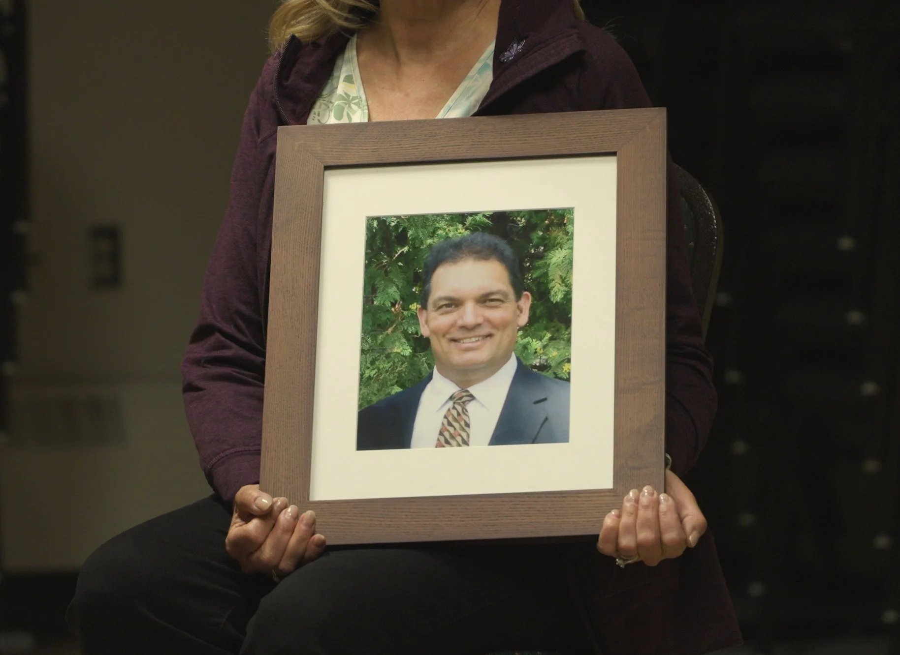 A woman holding a framed photo of a smiling man in a suit and tie, with green foliage in the background.