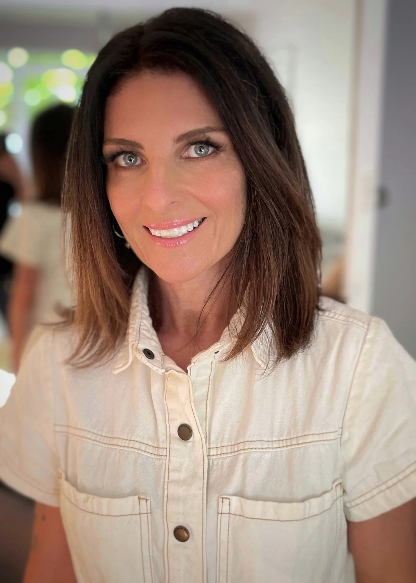 A woman with shoulder-length brown hair and blue eyes smiling at the camera, wearing a light-colored button-up shirt, standing indoors with blurred background.