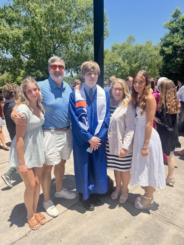 Group of people at graduation ceremony, with a young man in a blue cap and gown in the center, surrounded by four women and a man, all smiling outdoors on a sunny day.
