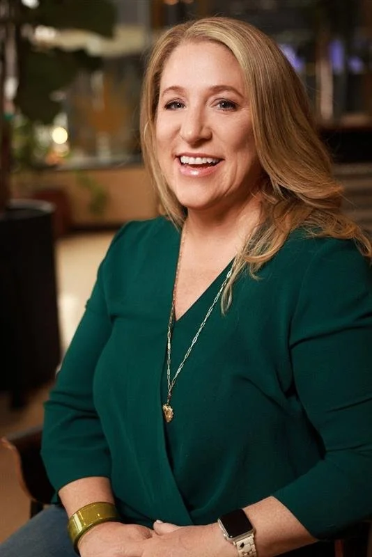 Confident woman with blonde hair smiling, wearing a green top, gold bracelet, silver necklace, and smartwatch, in an indoor setting.