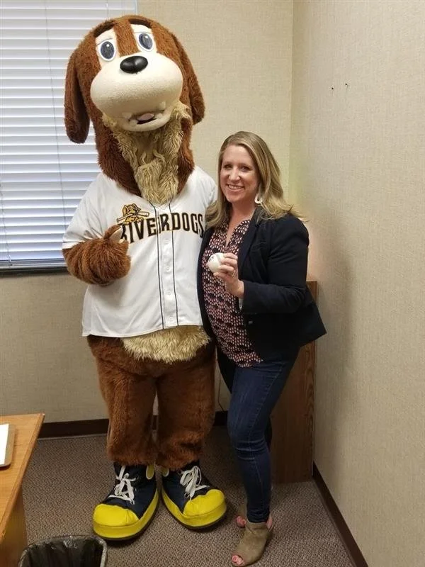 A woman standing next to a person in a dog mascot costume, both smiling. The mascot is wearing a white baseball jersey with 'Riverdogs' written on it and large yellow shoes. The woman is dressed in a dark blazer, patterned blouse, and jeans, holding a baseball in her hand.