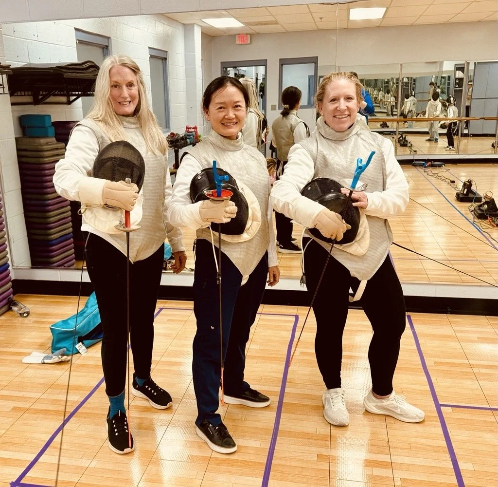 Three women in fencing gear holding fencing masks and foils, standing in a gym with mirrors and exercise equipment, smiling at the camera.