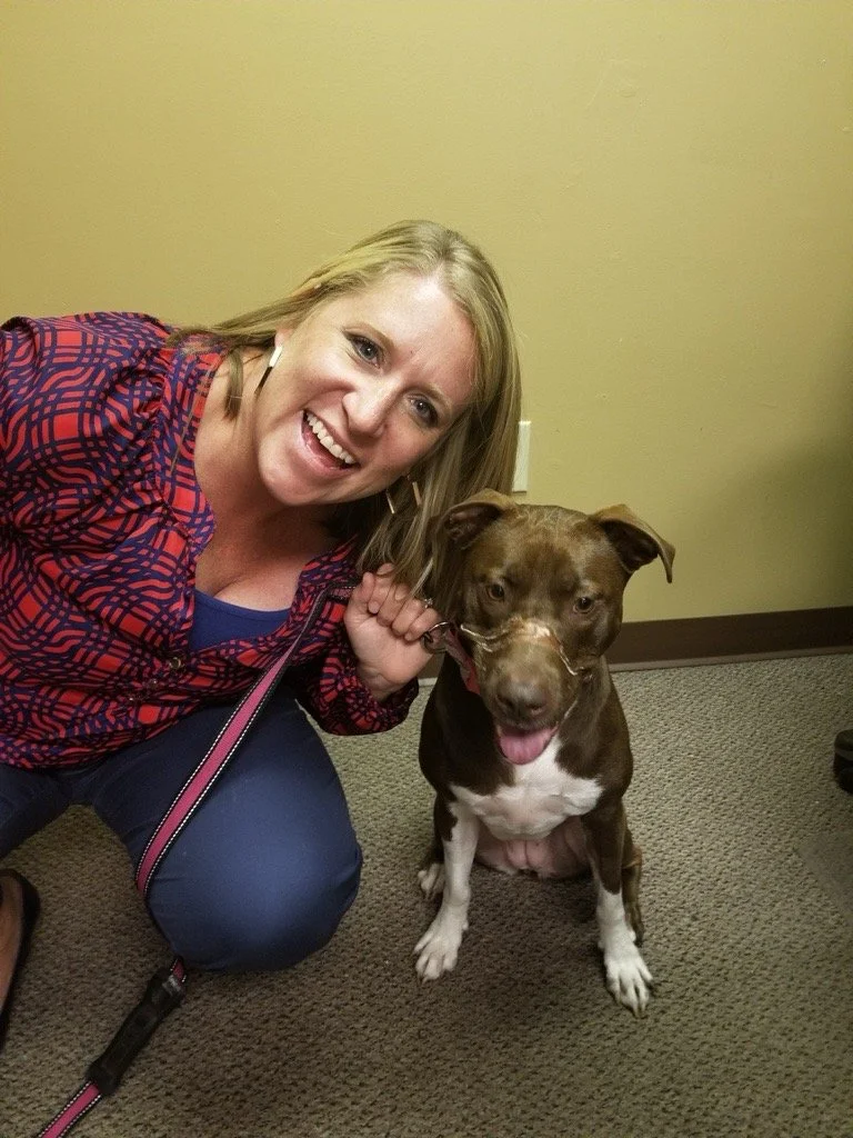 A smiling woman with blonde hair kneeling on carpeted floor next to a brown and white dog with a happy expression indoors.