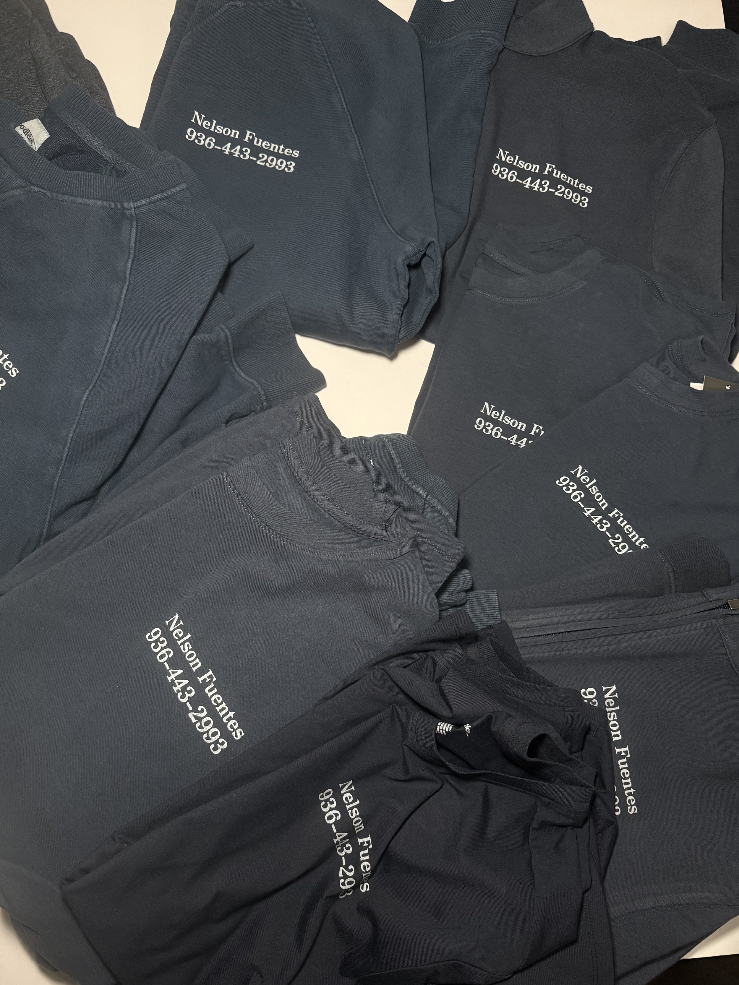 A collection of black and dark gray sweatshirts, T-shirts, and jackets with white text displaying the name "Nelson Fuentes" and a phone number "936-443-2993".