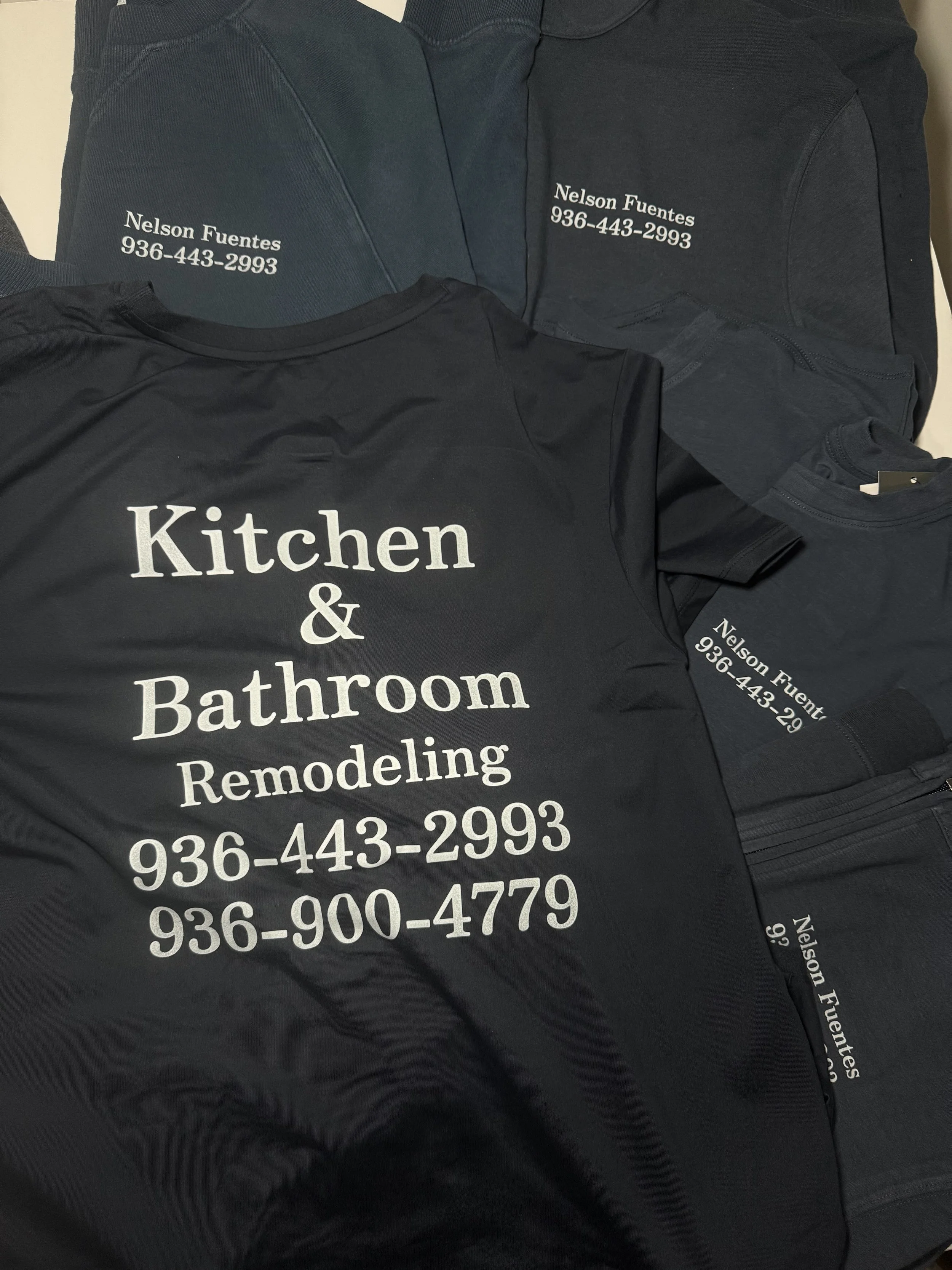 Black t-shirt with white text advertising kitchen and bathroom remodeling services and phone numbers, along with dark-colored sweatshirts with contact details embroidered on the chest.