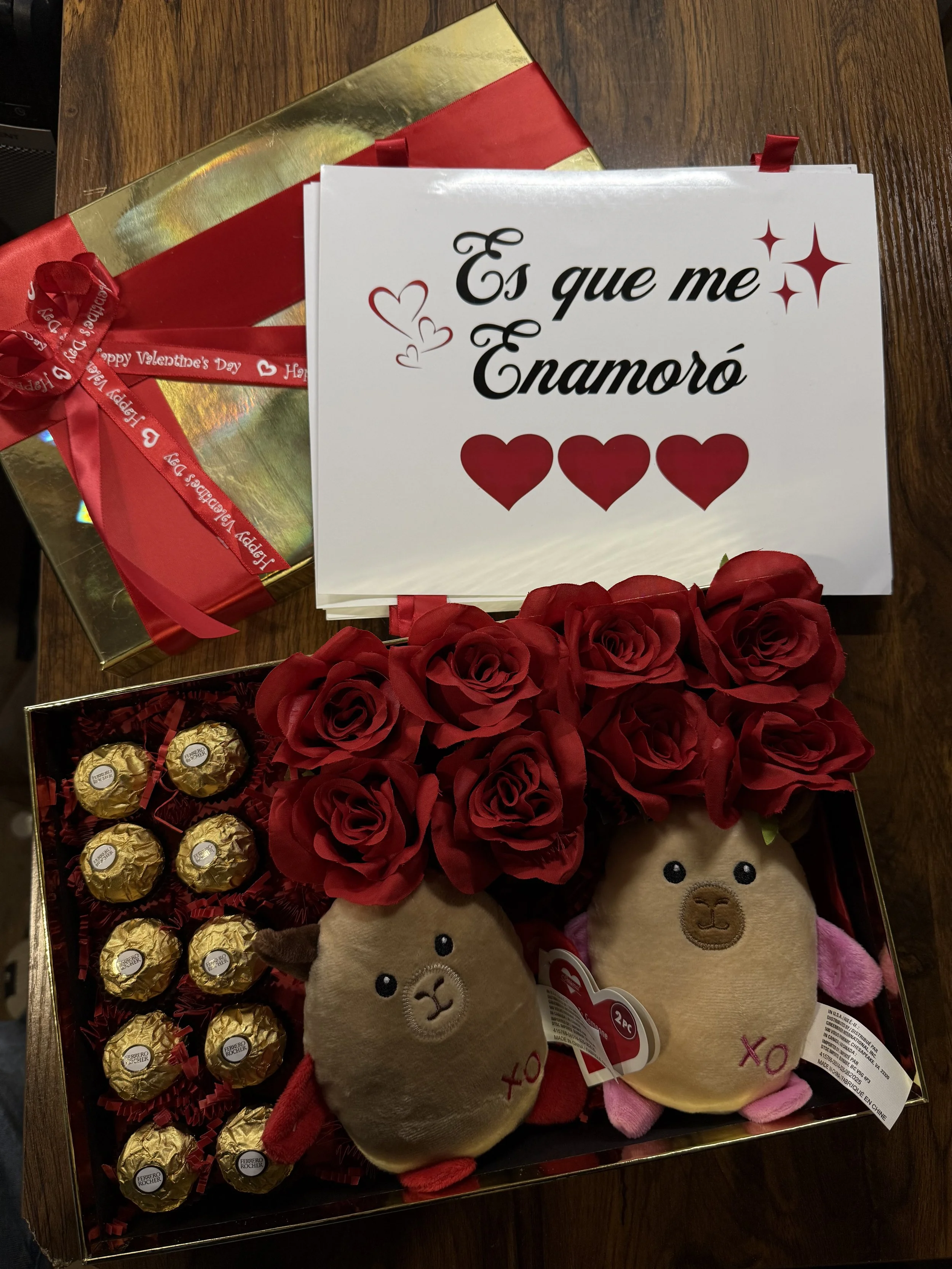 Valentine's Day gift set with stuffed animal toys, red roses, gold-wrapped chocolates, a white card with Spanish text, a gold gift box, and a red ribbon.