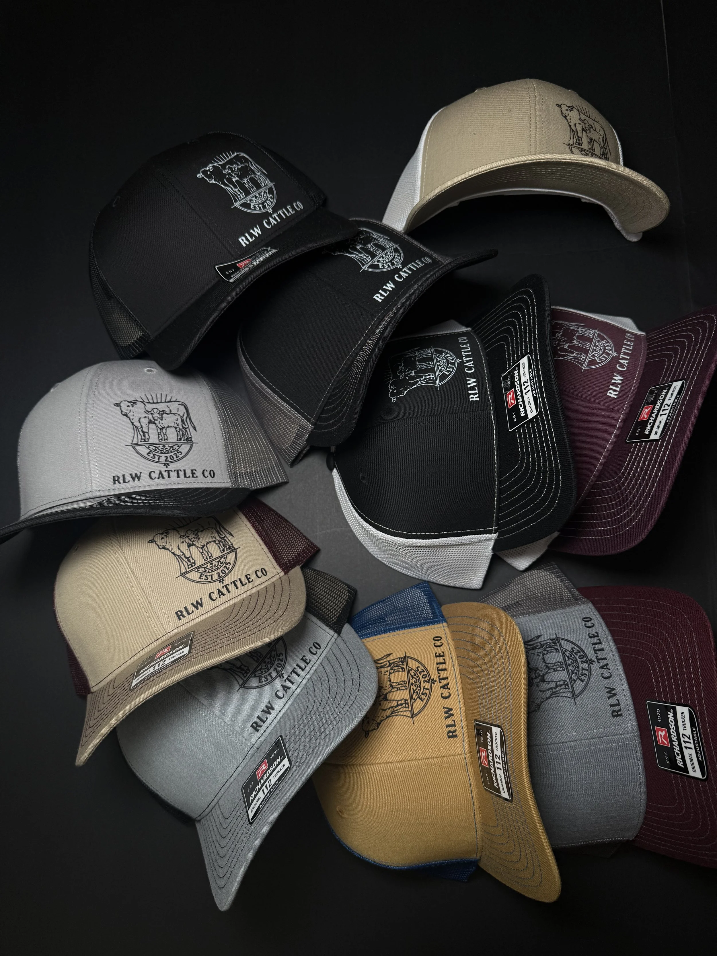 Collection of various colored baseball caps with 'RLW CATTLE CO' logo and emblem, arranged on a black surface.