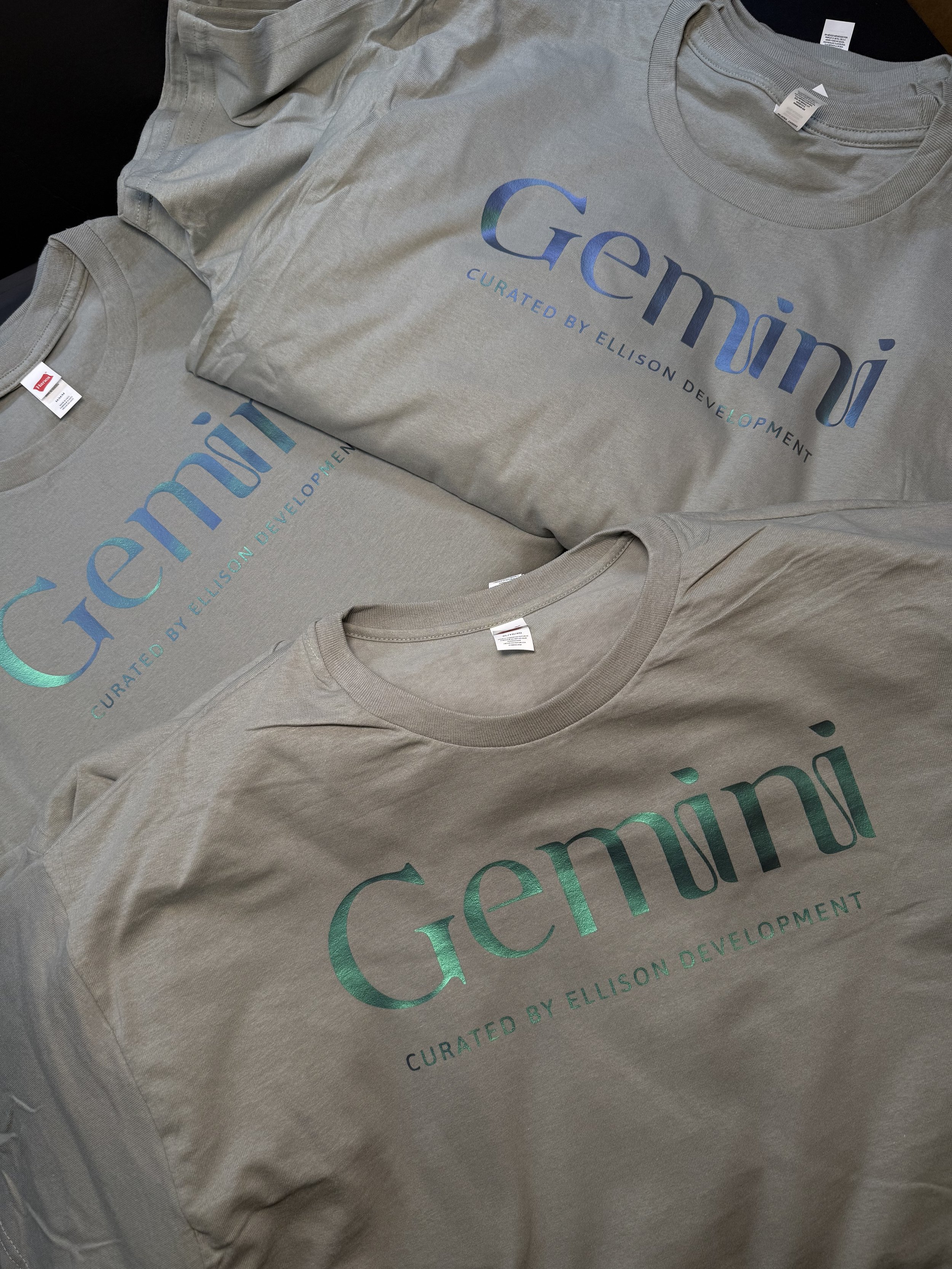 Three light gray T-shirts with the word "Gemini" and the phrase "Curated by Ellison Development" printed on the front in different shades of blue or green.
