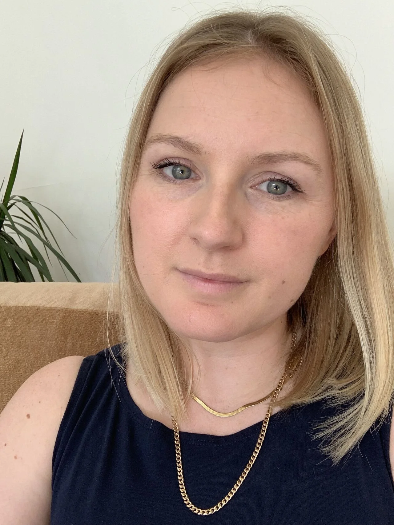 Lydia Wakefield-Yeung - A woman with shoulder-length blonde hair, blue eyes, wearing a navy sleeveless top, gold necklace, and sitting on a beige couch with a green plant in the background.