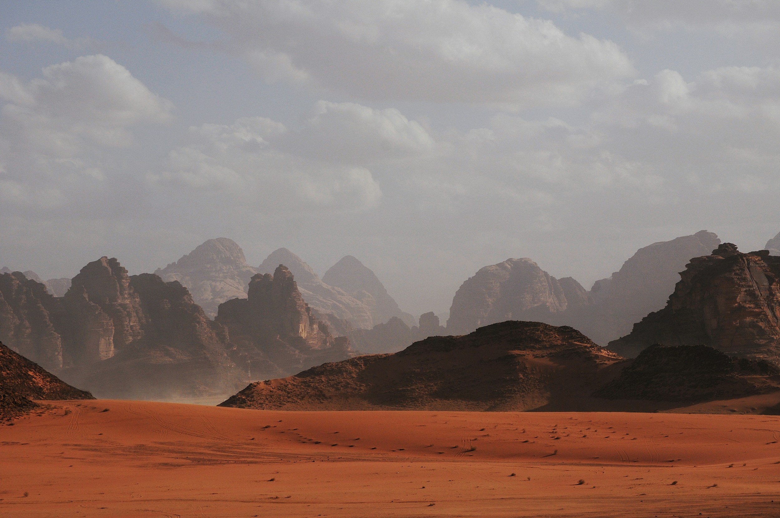 A desert landscape with reddish sand in the foreground, rugged mountains in the background, and a partly cloudy sky overhead.