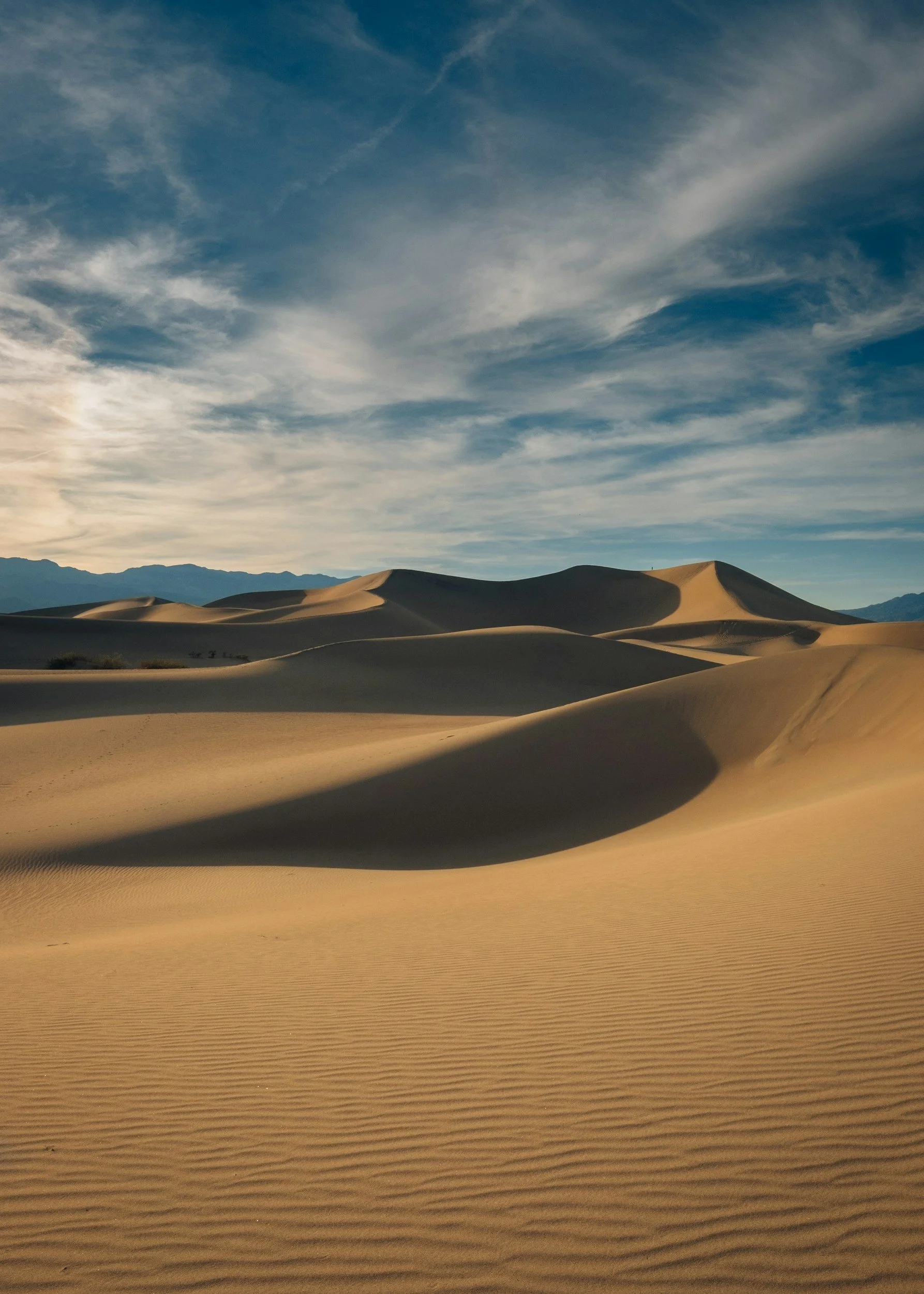 Sand dunes in a desert with a mountain range in the background and a partly cloudy sky.