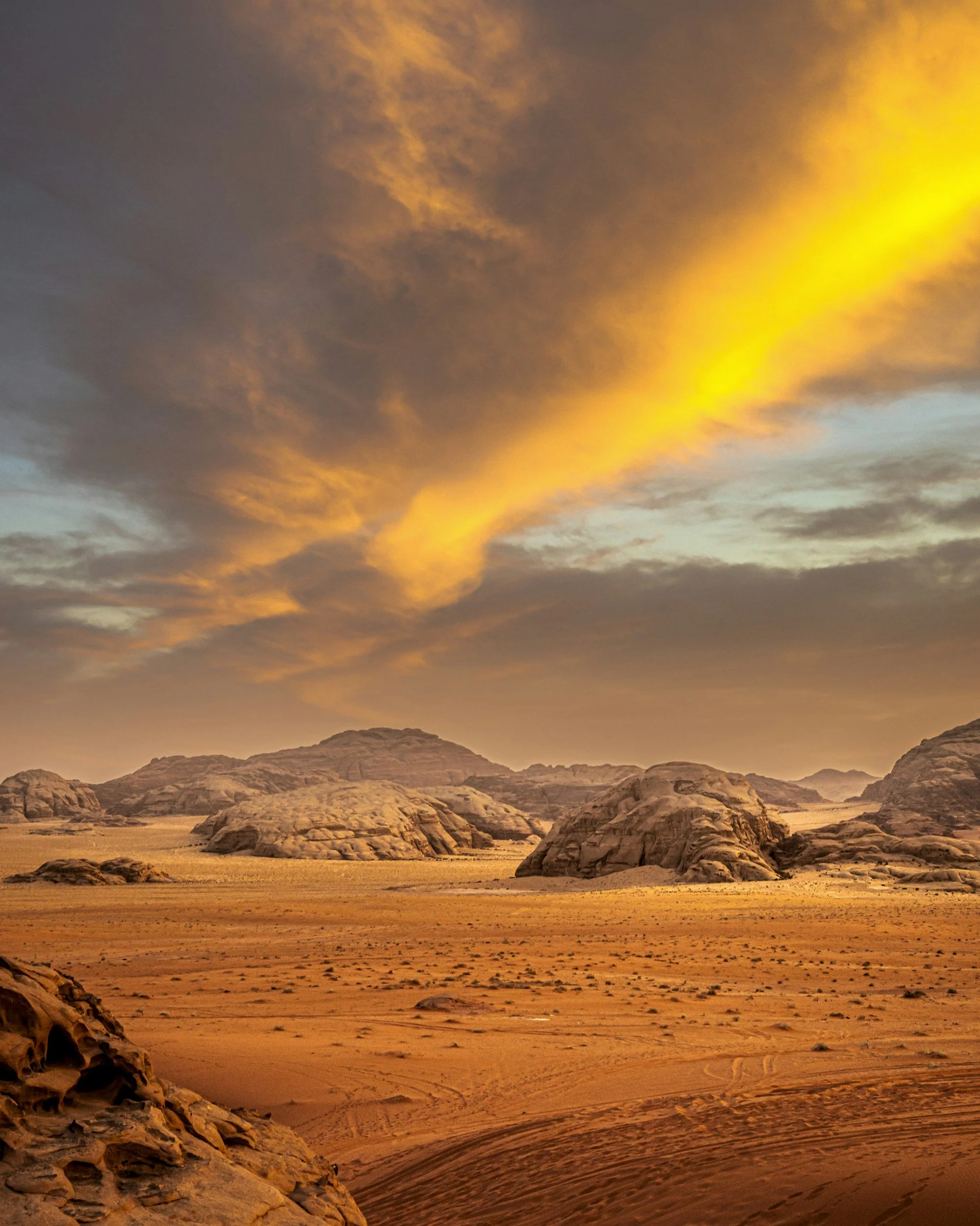 A desert landscape at sunset with rocky formations, sand dunes, and a dramatic sky with orange and yellow clouds.