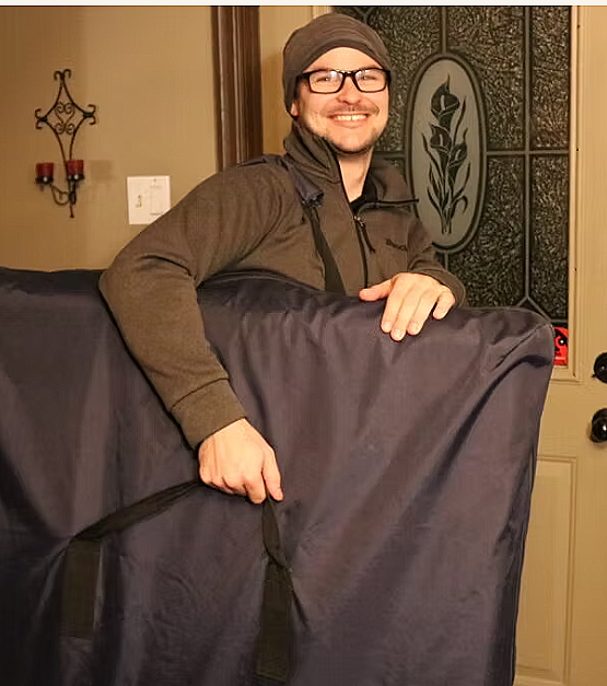 Un homme souriant portant des lunettes, un bonnet et un manteau, tenant une grande couverture ou un sac de voyage dans une pièce domestique.