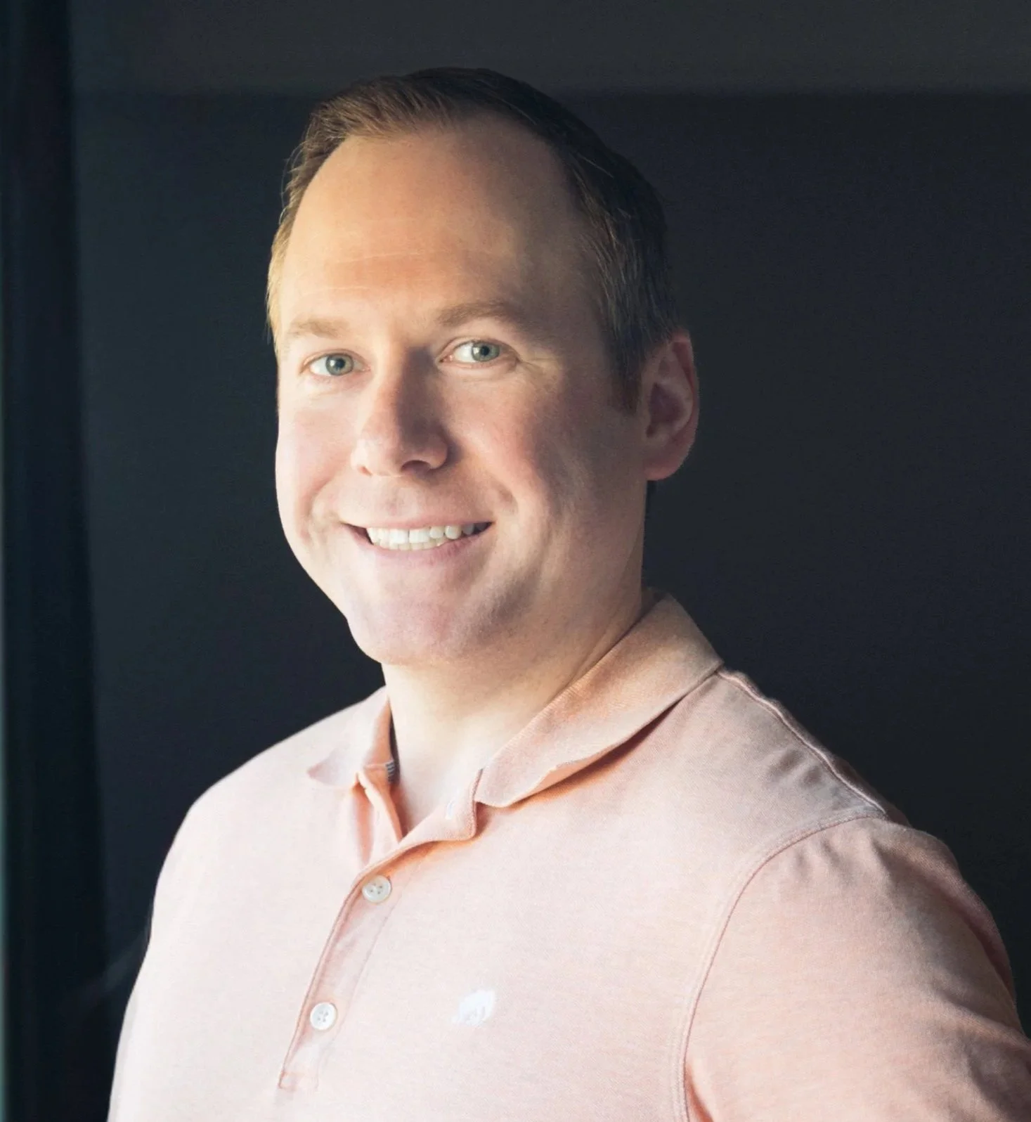 Portrait of a smiling man with light-colored hair, blue eyes, wearing a peach-colored button-up shirt, standing against a dark background.