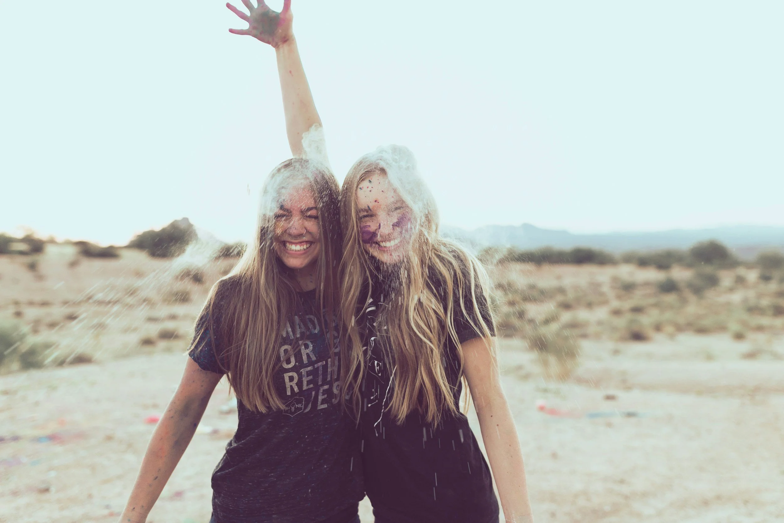 Two young women smiling and standing close together as water splash hits them in an outdoor desert landscape.