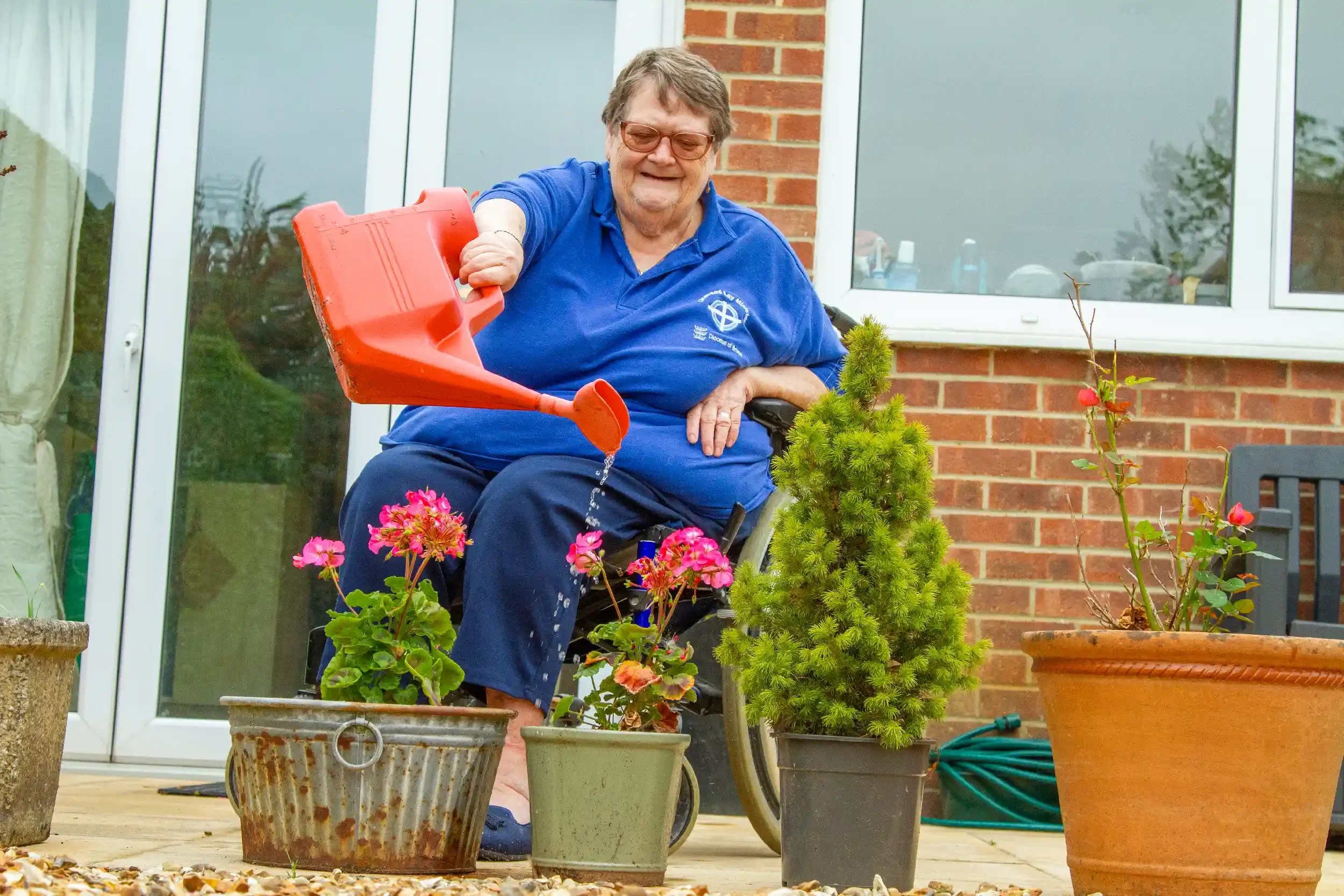 An elderly woman in a wheelchair watering potted plants outside a brick house.