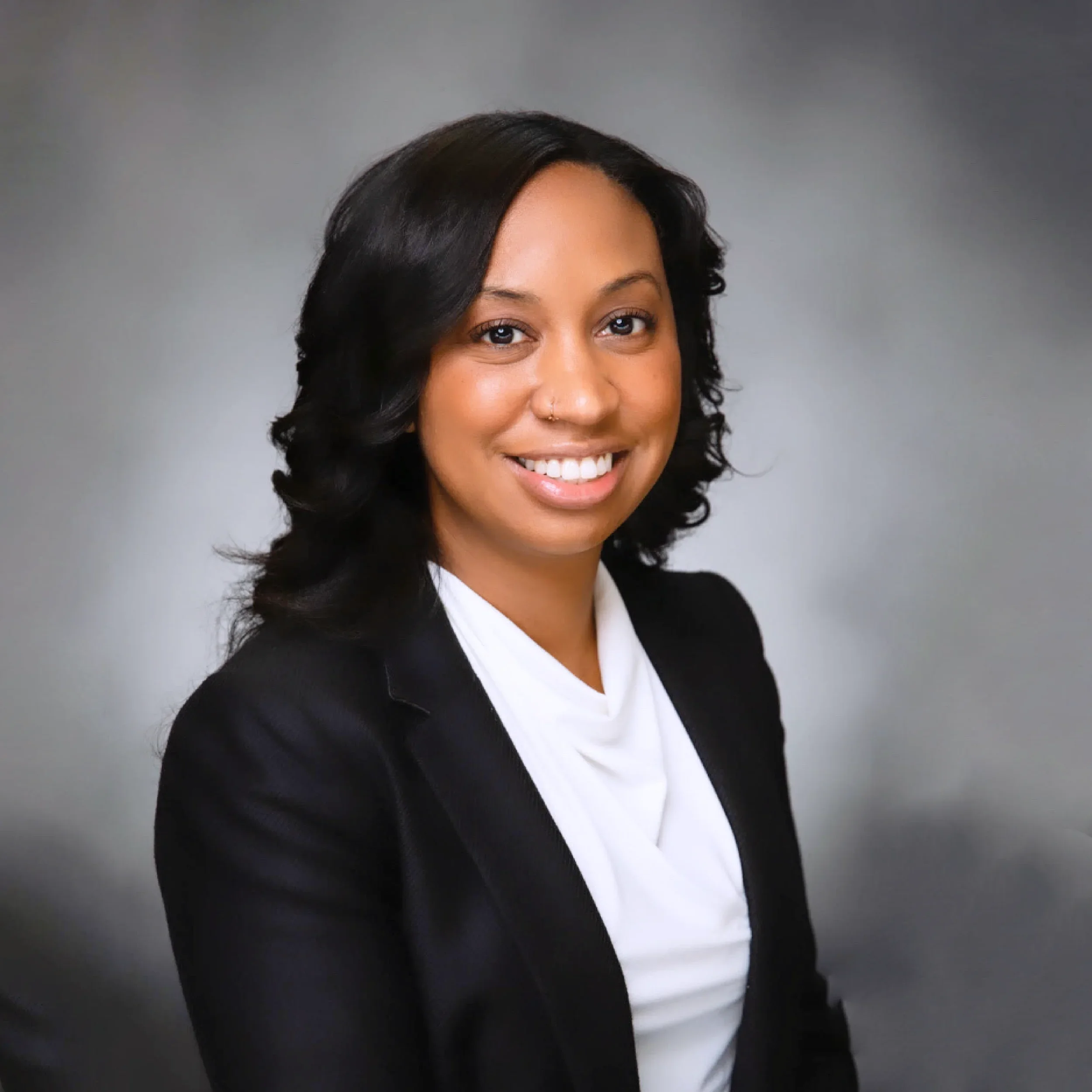 Professional headshot of a woman with black hair, smiling, wearing a black blazer and white blouse against a gray background.