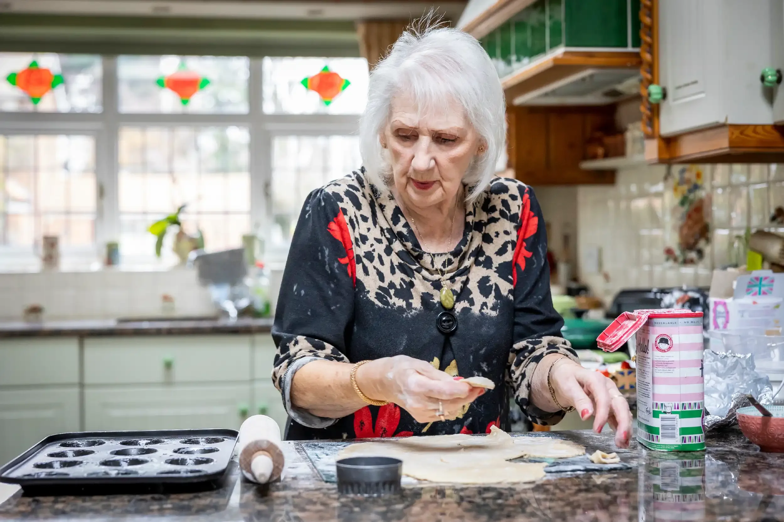 An elderly woman with white hair wearing a black and red patterned top is baking in a kitchen, rolling out dough with a rolling pin nearby, surrounded by baking supplies.