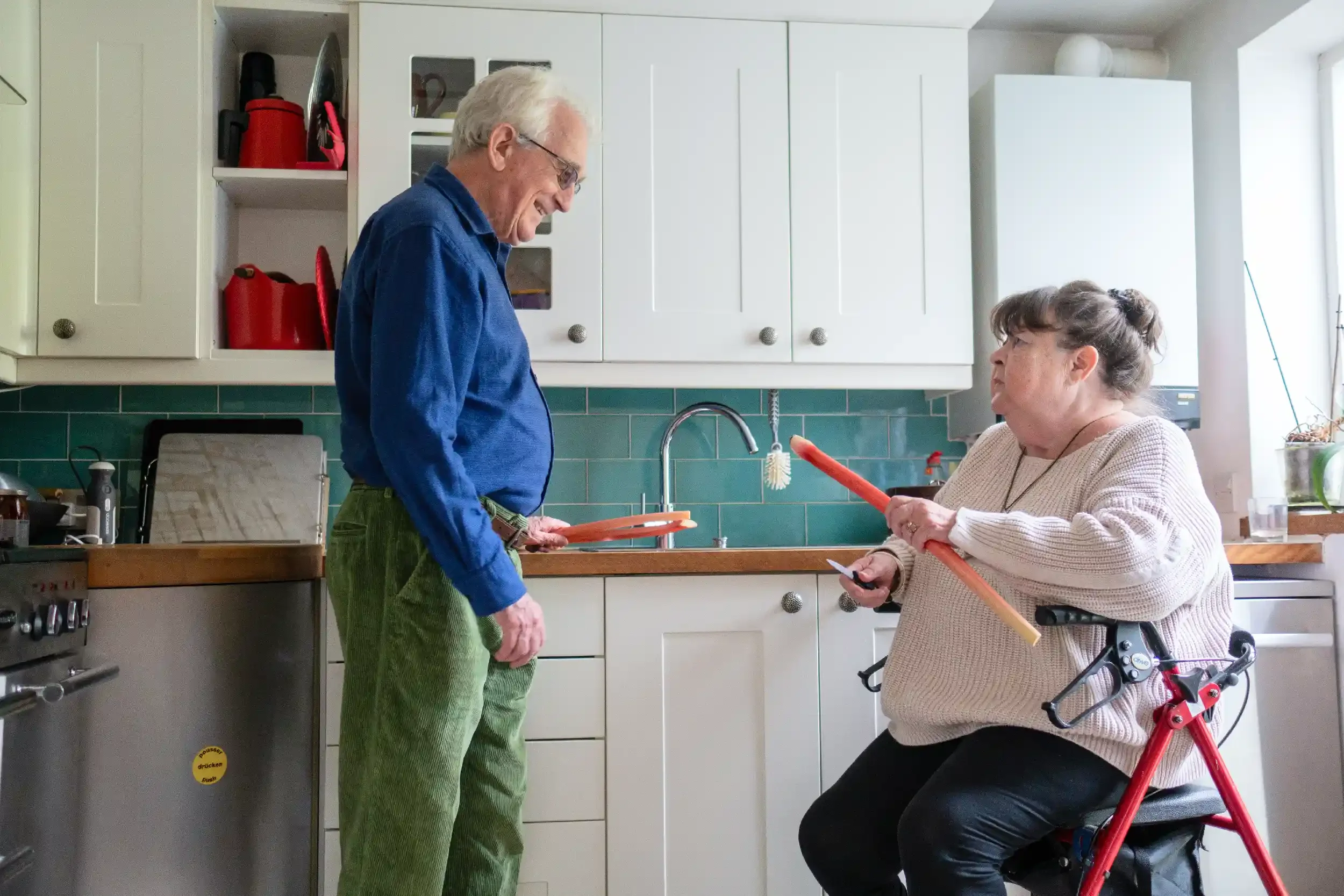 A man and a woman are in a kitchen, holding orange broomsticks and smiling at each other. The woman, sitting in a red mobility chair, is holding a remote control. The man is looking down at her, and the kitchen has white cabinets, a green tiled backsplash, and a window letting in natural light.