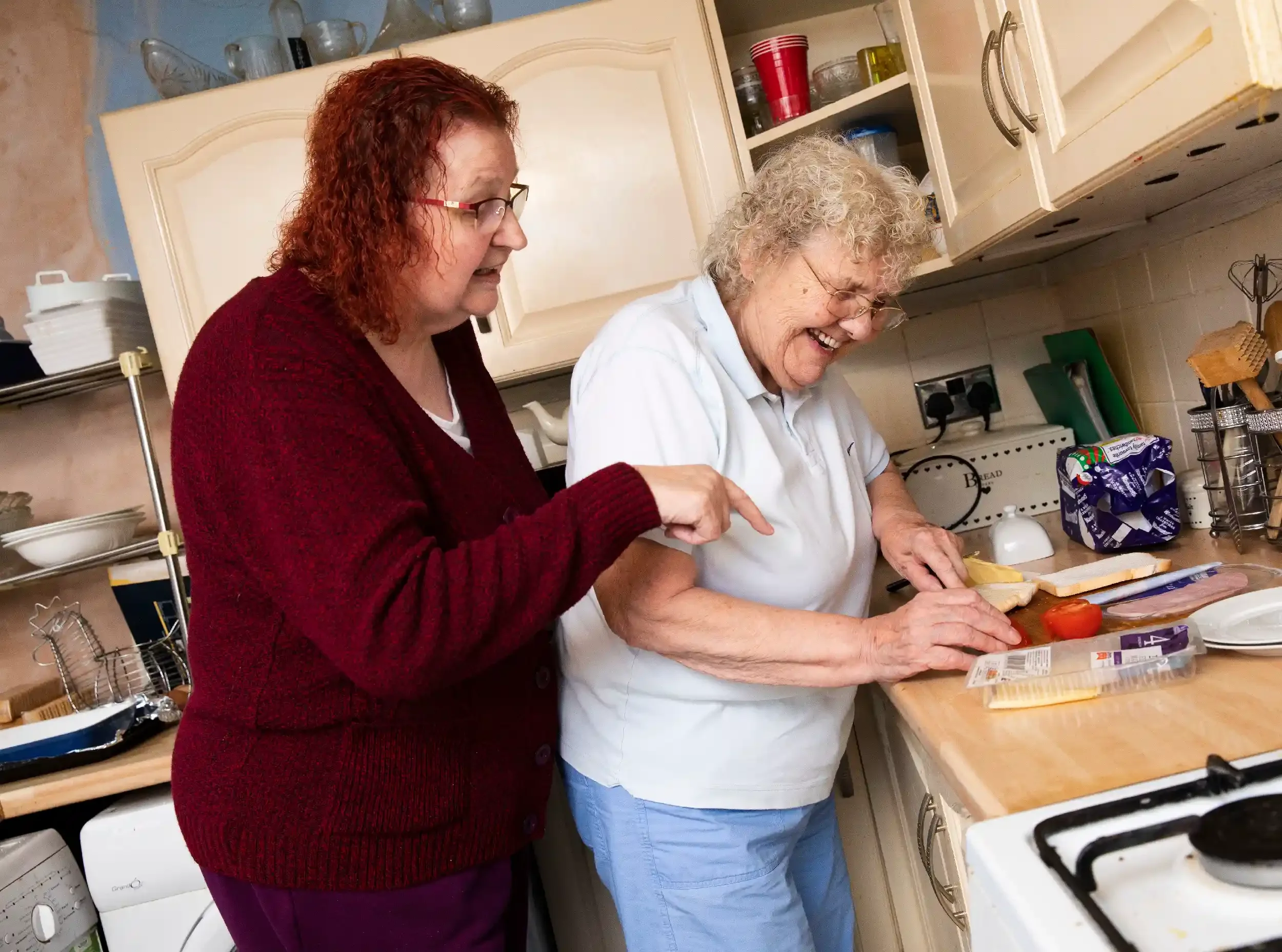 Two women are preparing sandwiches together in a kitchen, laughing and smiling.
