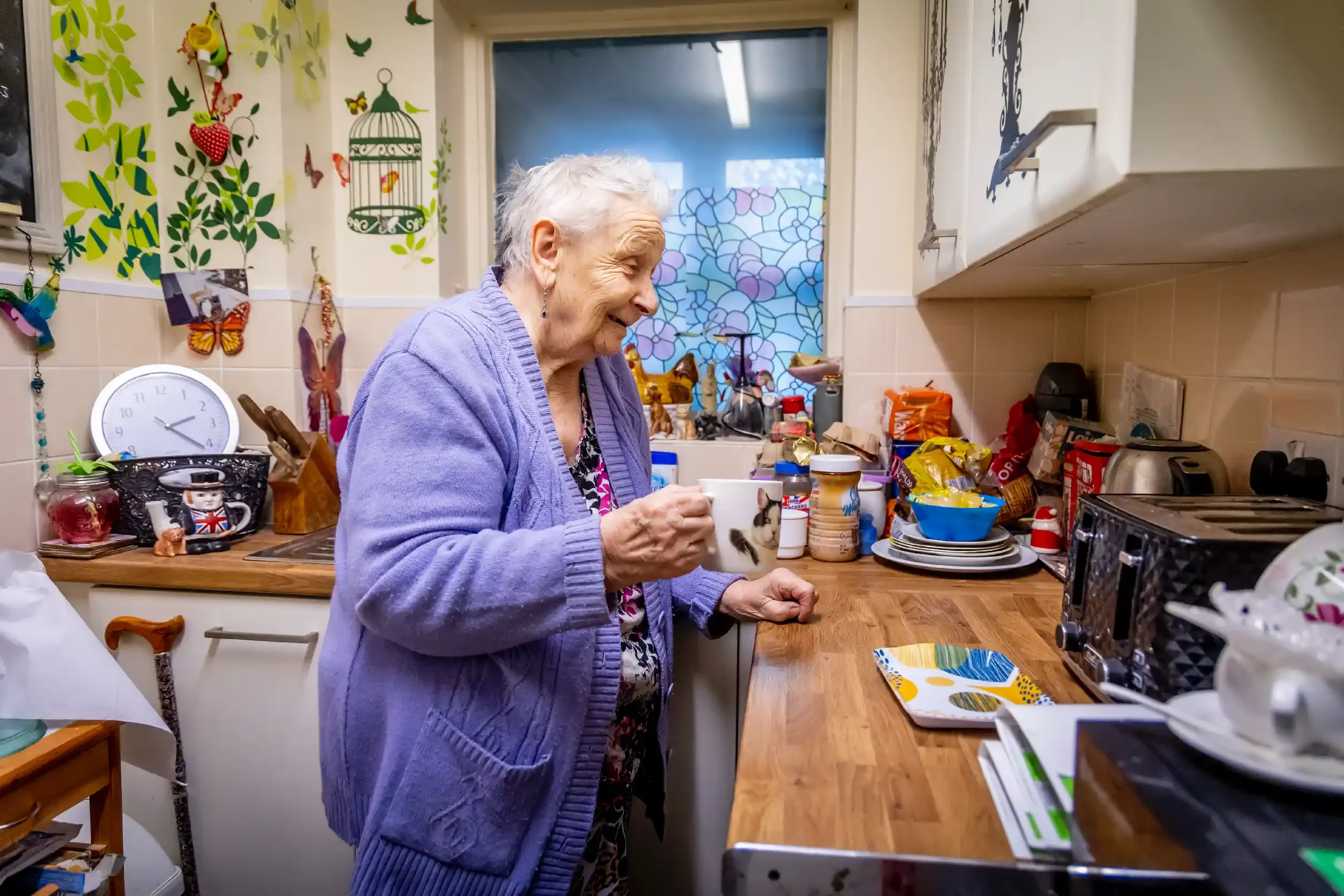 An elderly woman with gray hair, wearing a purple cardigan, stands in a cluttered kitchen, holding a mug, smiling.