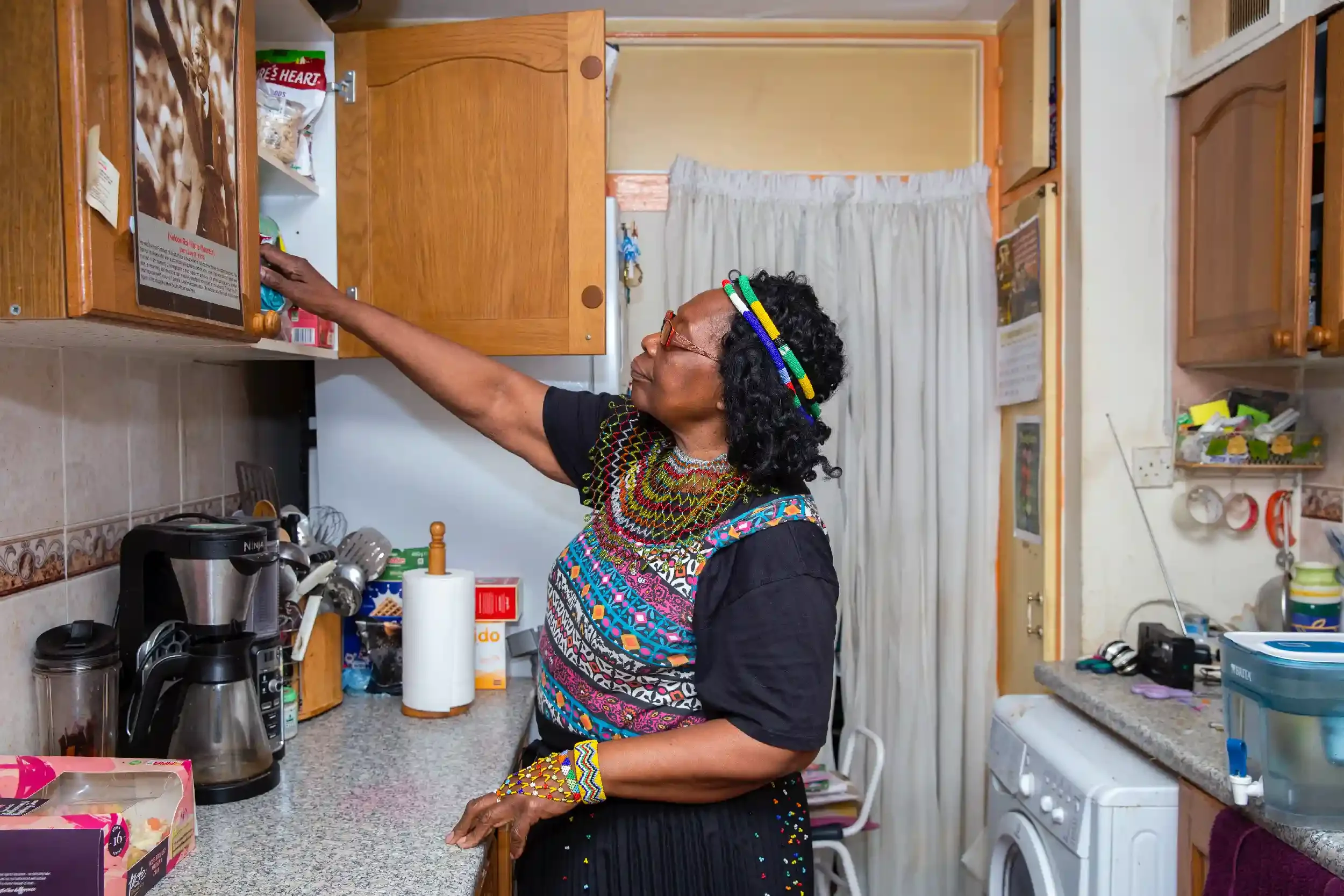 A woman with colorful beaded jewelry and a beaded headband reaches into a kitchen cabinet with wooden cabinets, a coffee maker, and various kitchen items around her.