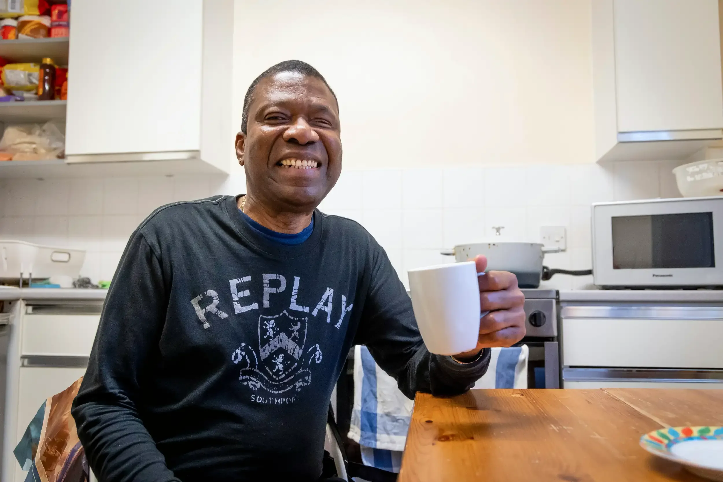 A man sitting at a kitchen table holding a white mug, smiling, with a stove, microwave, and kitchen shelves in the background.