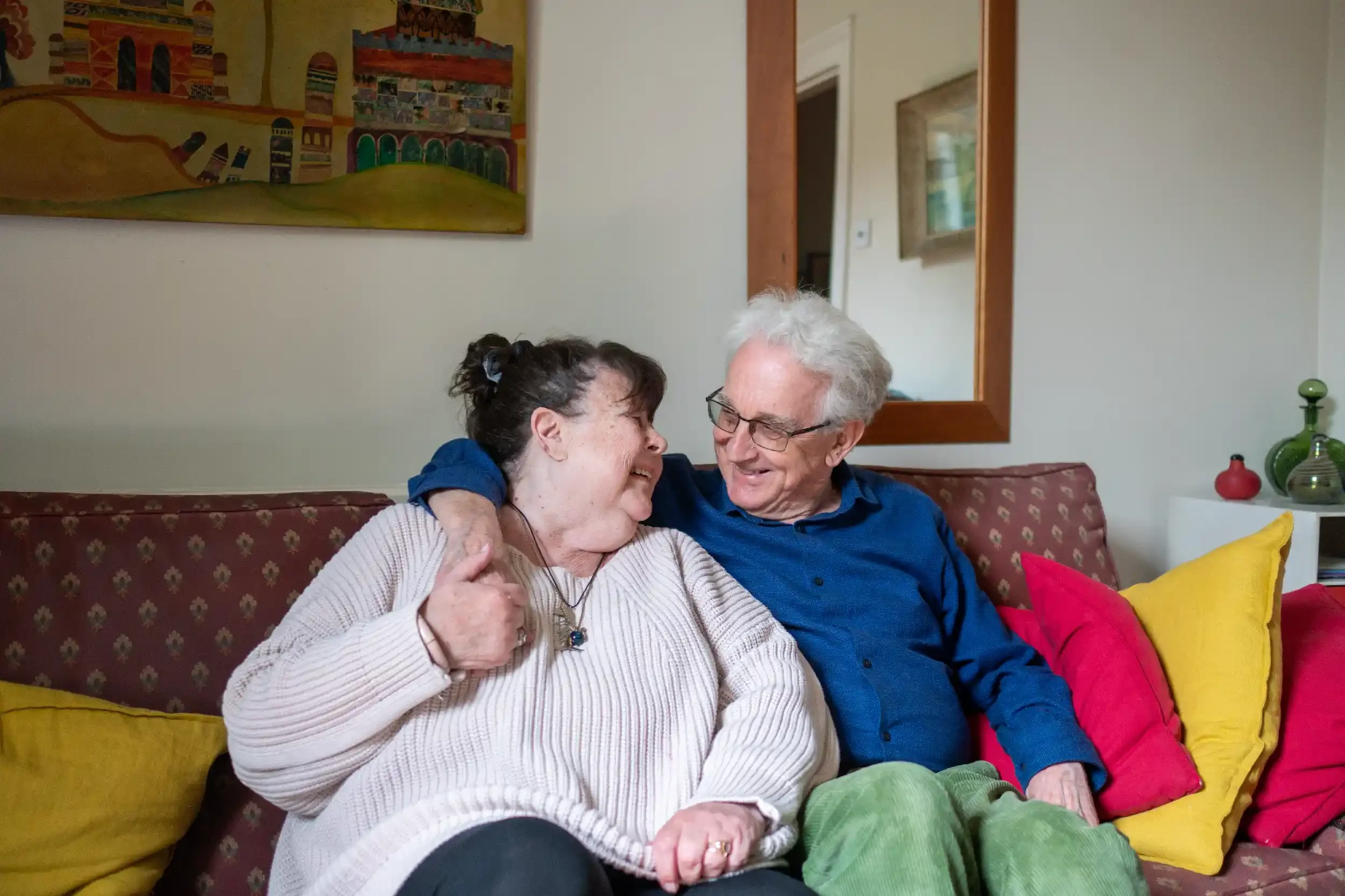 An elderly couple sits on a couch, smiling and laughing together, sharing an intimate moment in a cozy living room with colorful pillows and artwork on the wall.